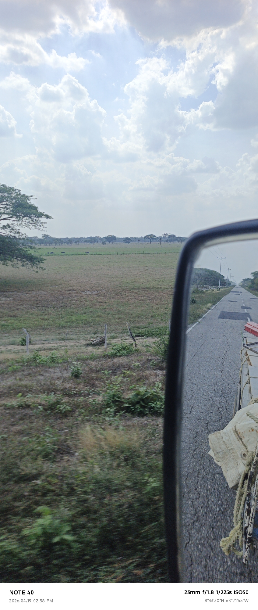 This is a slightly blurry, eye-level, daytime shot taken from a moving vehicle in what appears to be a rural area in Arismendi, Venezuela. The sky is bright with scattered white clouds. To the left of the frame is a field of tall grass with a barbed wire fence. Beyond that, there's a line of trees in the distance. To the right, part of the vehicle's side mirror is visible, along with a glimpse of the road and what seems to be the back of a truck bed loaded with cargo and tied down with ropes. The GPS coordinates are 8°33'30"N, 68°21'45"W. The camera settings are 23mm, f/1.8, 1/225s, ISO50.
