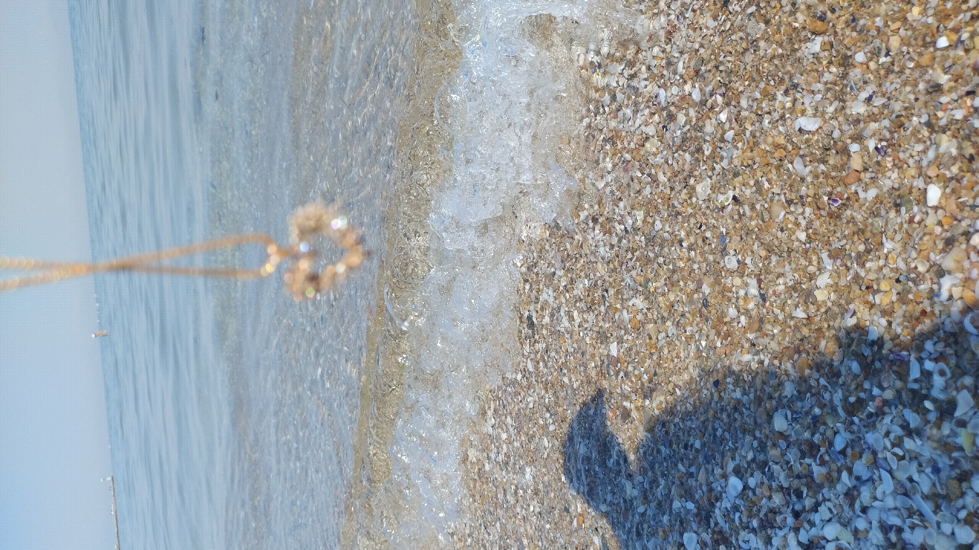 The image depicts a close-up view of a shell-covered beach with the sea in the background. A delicate, gold-colored heart-shaped pendant with small, clear stones is suspended by a thin chain, hanging into the frame from the top. The pendant is slightly out of focus. A gentle wave is breaking on the shore, creating a foamy, translucent curtain of water that washes over the shells. The beach is densely covered with a variety of small shells and pebbles in shades of white, beige, brown, and gray. In the lower left portion of the image, a dark shadow is cast on the shells, suggesting the presence of a person or object out of frame, to the left. The water in the background is calm and appears a light blue, with faint ripples. In the distance, on the horizon, two indistinct figures or structures are visible in the water, possibly buoys or distant people. The overall scene suggests a peaceful, sunny day at the beach.