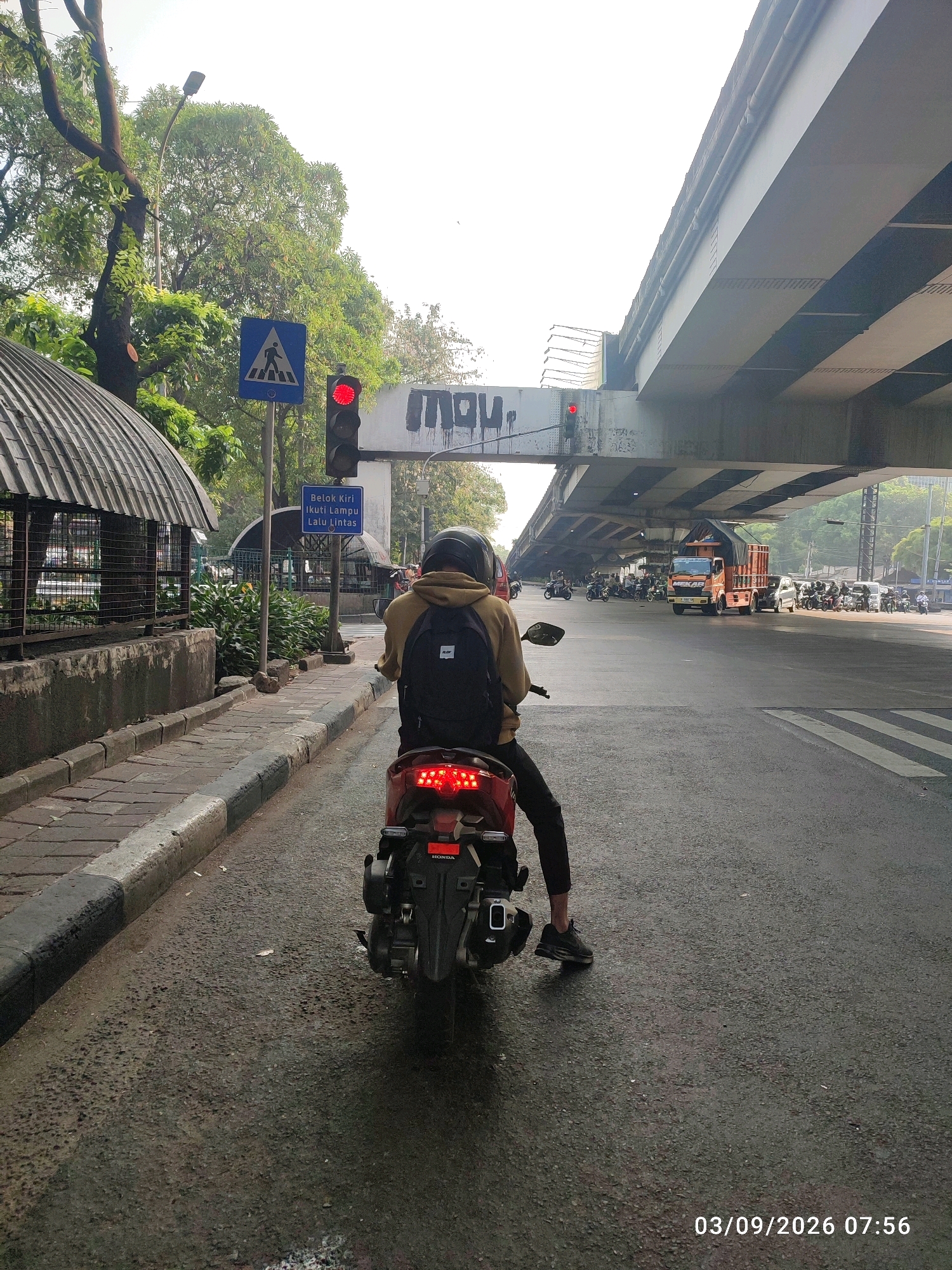 The image captures a street scene in East Jakarta, Indonesia, likely in the vicinity of the Cililitan Overpass, a prominent flyover that facilitates traffic flow. A person wearing a tan hoodie and black pants rides a red motorcycle with a black backpack. The motorcycle is positioned on the road, facing away from the viewer and towards the overpass. The traffic light is red, indicating that the motorcycle is stopped.
The environment includes a gray concrete overpass spanning overhead. Below the overpass, several other vehicles are visible, including a bright orange truck with a cargo bed, a white car, and numerous motorcycles. The road is paved with asphalt, showing signs of wear and some dampness. To the left of the road, there's a sidewalk made of gray paving stones with a black curb. A fence with a curved, ribbed metal roof structure is also present on the left side of the frame, possibly part of a building or an enclosed area. Lush green trees with dense foliage line the background.
A blue pedestrian crossing sign with a white silhouette of a person walking is mounted on a pole, positioned to the left of the traffic light. Below the pedestrian sign is another blue sign with Indonesian text that reads "Belok Kiri Ikuti Lampu Lalu Lintas," which translates to "Turn Left Follow Traffic Lights." Graffiti is visible on the side of the overpass structure, including the word "inay" in black spray paint. The date and time stamp at the bottom right corner indicates "03/09/2026 07:56." The scene is illuminated by natural daylight, suggesting it is morning.