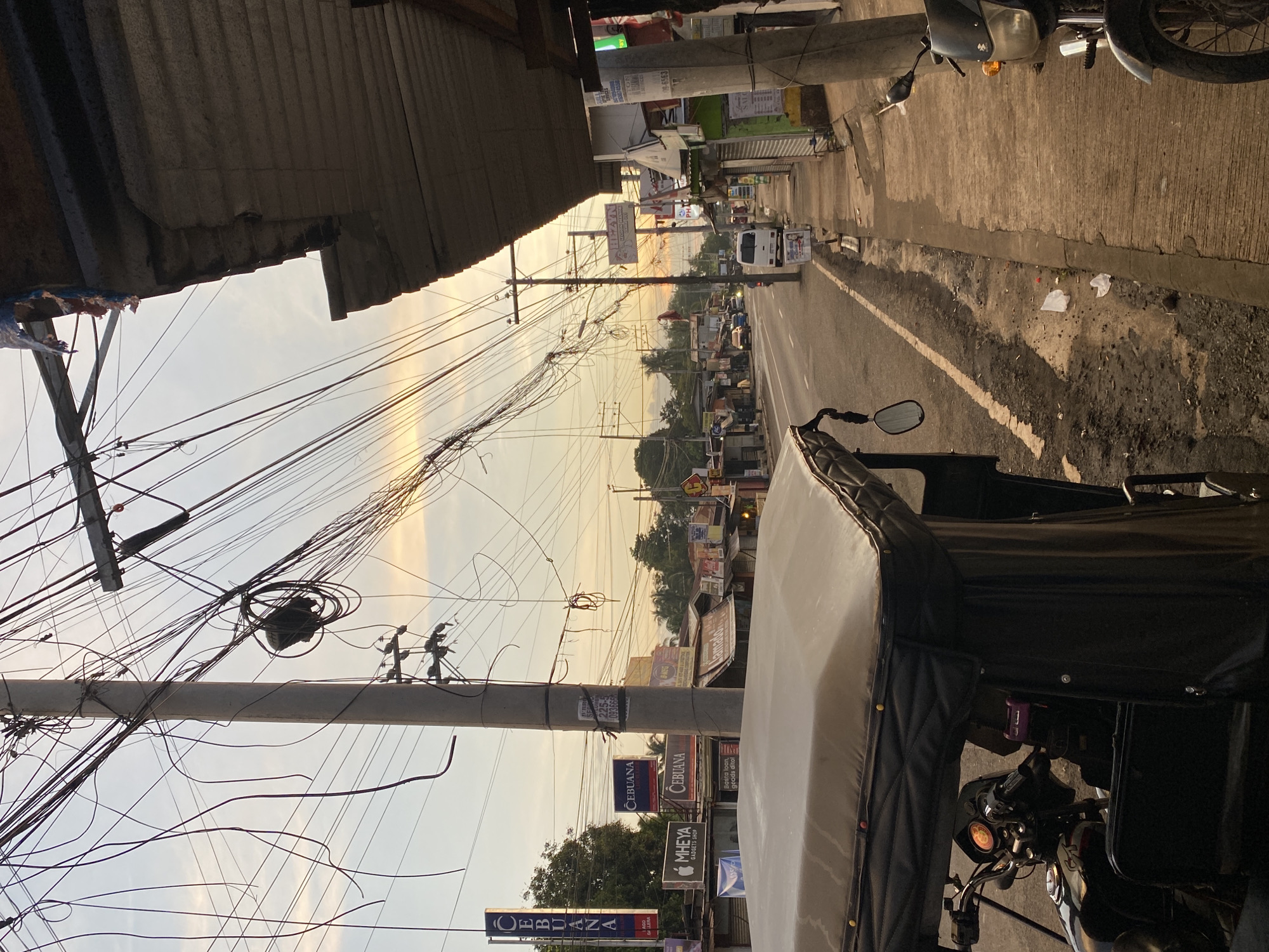 The image depicts a street scene in Davao City, Philippines, during what appears to be sunset or sunrise. The foreground features the rear of a three-wheeled motorized vehicle, likely a tricycle, with its dark fabric canopy and a portion of its body visible. To the right, the front of a black motorcycle is seen, parked on a concrete sidewalk. The sidewalk is lined with a variety of small businesses, indicated by signs such as "CEBUANA LHUILLIER," "MHEYA GADGETS SHOP," and other unreadable signage. Many of these businesses have corrugated metal roofs. A prominent utility pole is situated near the center of the image, heavily laden with a complex network of tangled electrical and telecommunication wires. The street itself is paved and appears to be a main thoroughfare, with a few vehicles, including a white van, visible in the distance. The sky displays warm hues of orange and yellow, suggesting either dawn or dusk. The overall environment appears to be a typical urban or semi-urban setting in the Philippines, characterized by dense overhead wiring and a mix of commercial establishments.