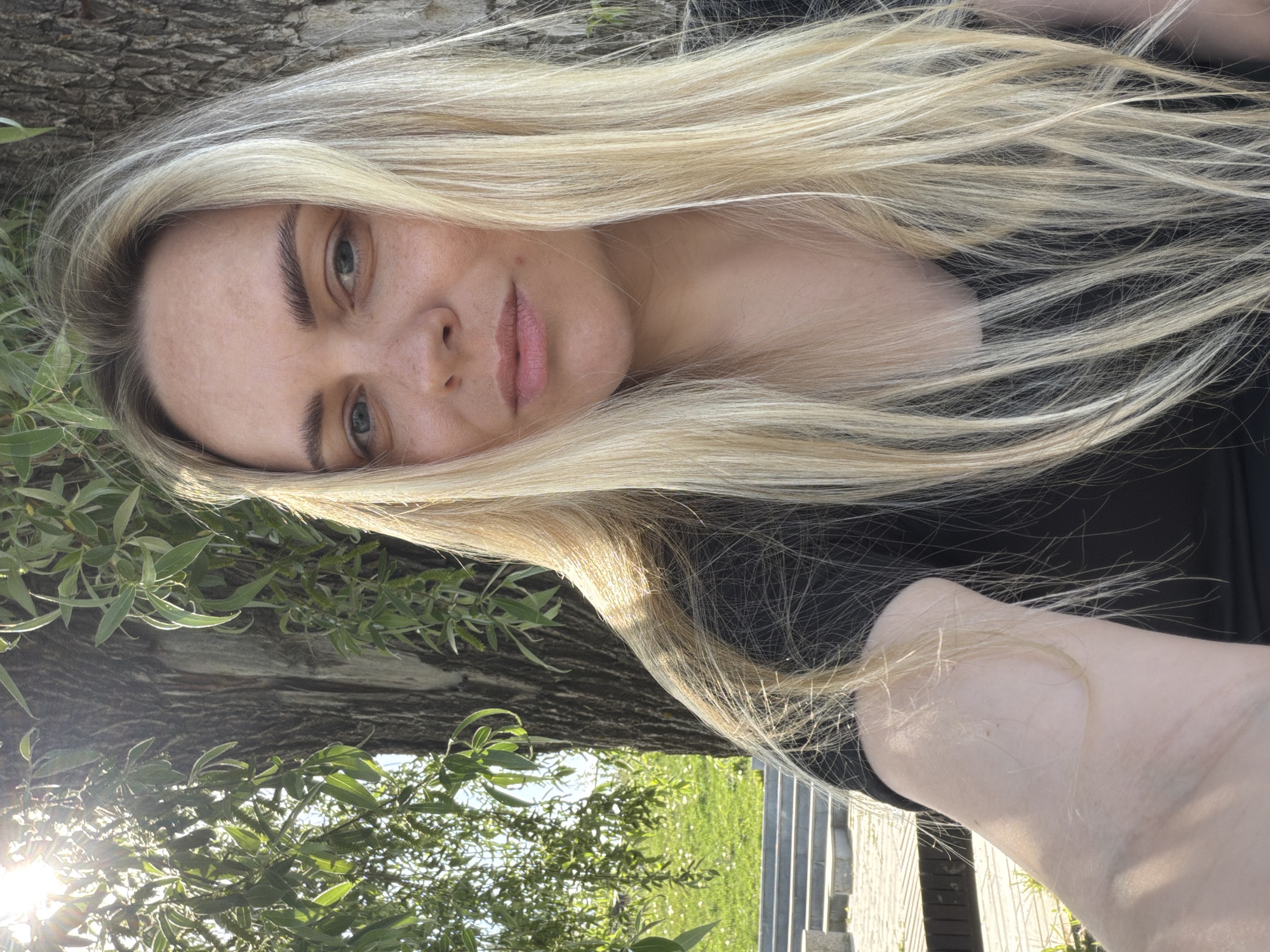 A woman with long blonde hair is positioned with a tree trunk behind her. She has fair skin, light eyes, and pink lips. She is wearing a black top. Outside, there are green leaves on branches, possibly from the tree. There appears to be sunlight filtering through the leaves. The location is Votkinsk, Russia.