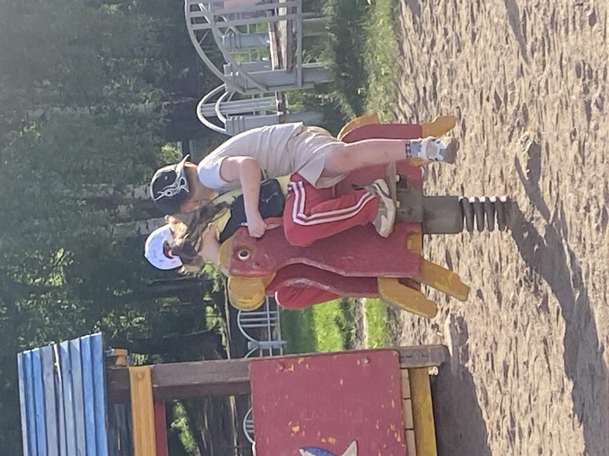 The image shows two children riding a red and yellow spring rider shaped like a bird in a playground. One child is wearing a white shirt and khaki shorts with a black baseball cap, and is facing forward. The other child is wearing a white helmet and red pants with white stripes. The playground is located in Chaikovsky, Russia, as evidenced by geotagging data. In the background, there is a metal climbing structure, trees, and sand. A wooden fence is visible in the foreground.