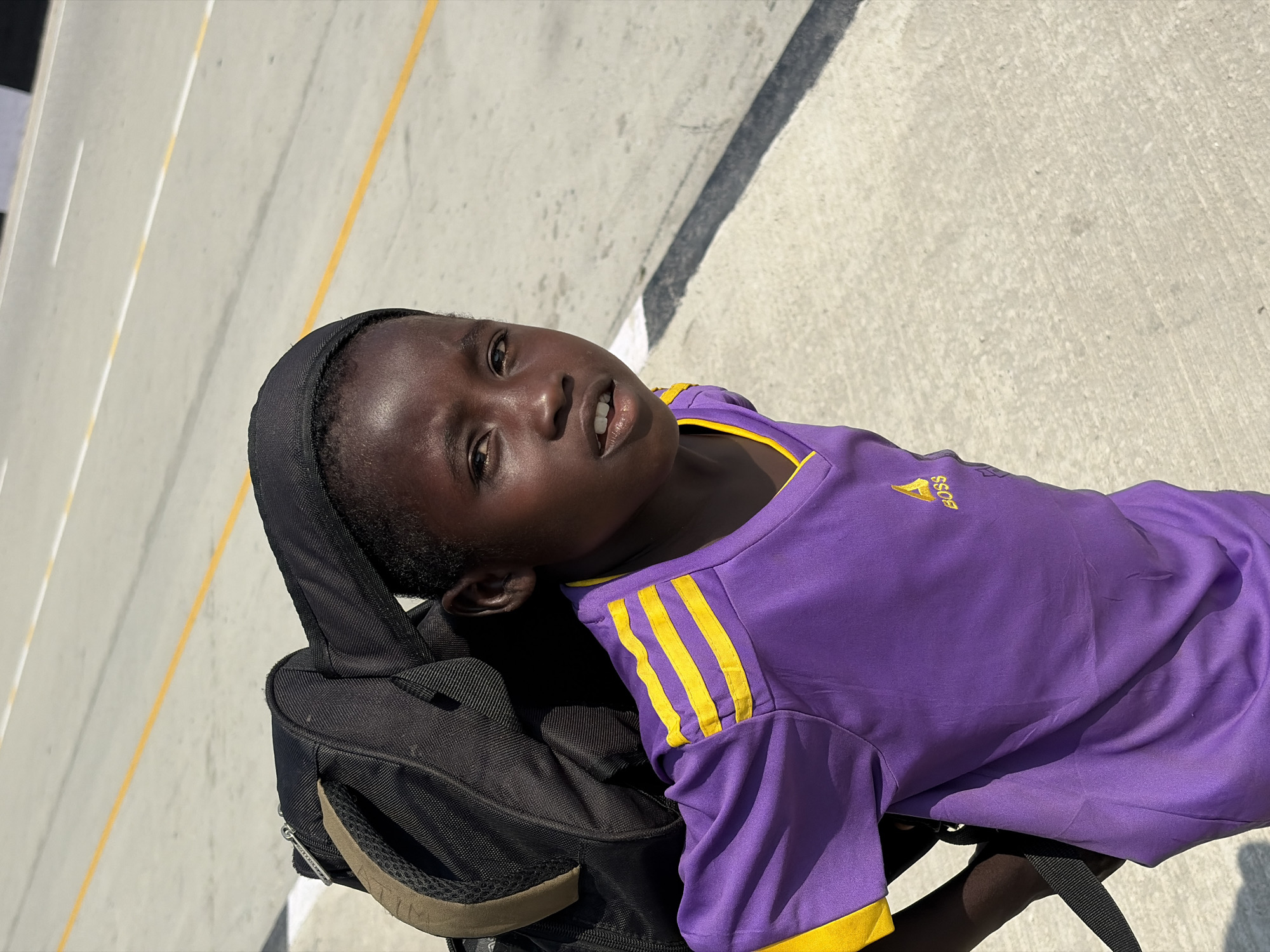 A young person stands in Alaguntan, Nigeria. The individual wears a purple shirt with yellow stripes on the shoulders and the word "Boss" embroidered on the chest and a black head covering. The individual carries a black backpack on their back. The setting appears to be an outdoor paved area with yellow and white lane markings.