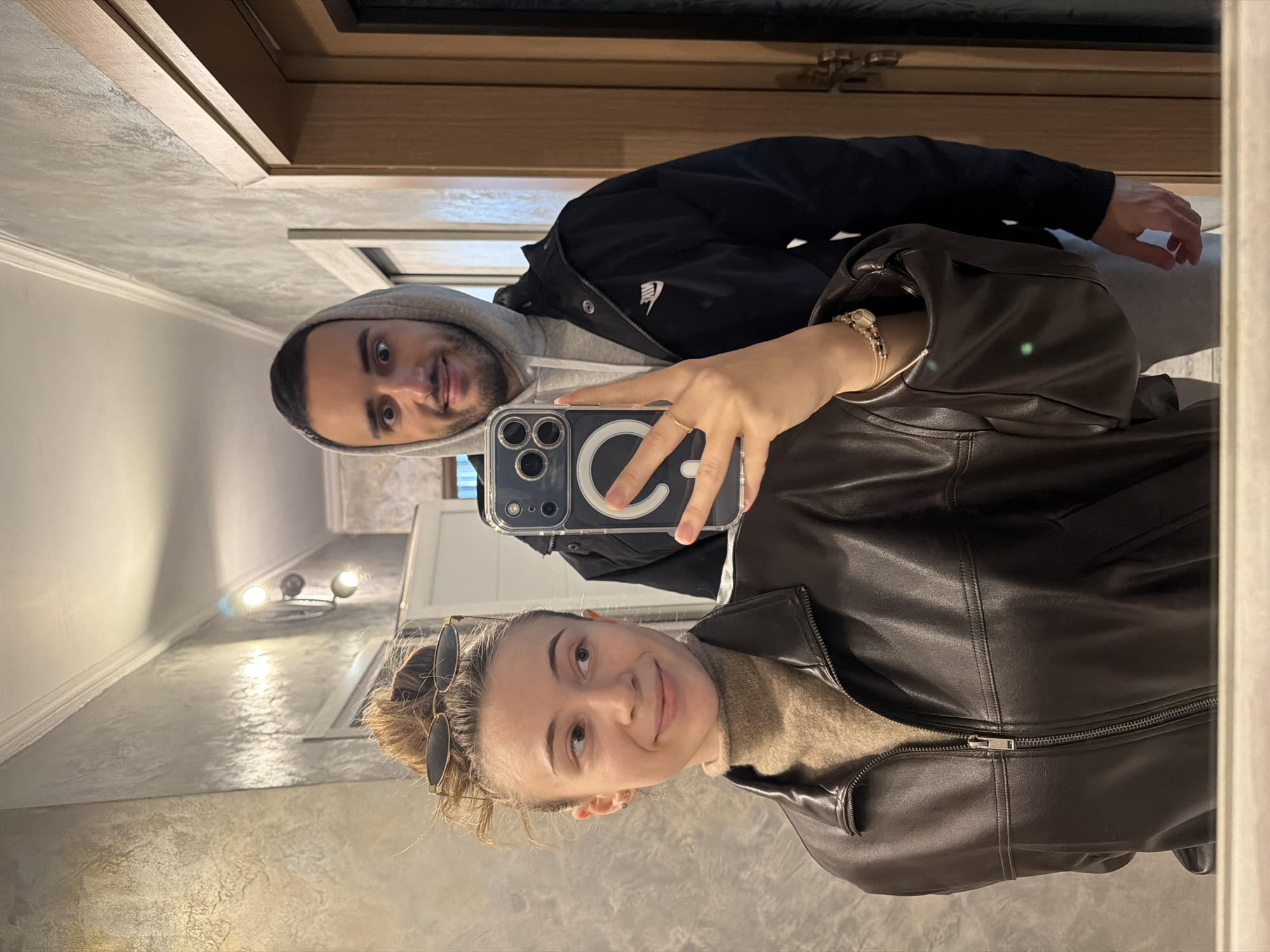 An indoor mirror selfie captures two individuals, a female in the foreground and a male behind her, located within a building in Plovdiv, Bulgaria. The female, positioned closer to the mirror, is seen from the chest up, looking to her right with a slight smile. She has light brown hair tied in a bun atop her head, with dark-rimmed sunglasses perched on top. She wears a beige turtleneck sweater underneath an unzipped dark brown leather jacket. A gold ring is visible on her left ring finger, and multiple beaded bracelets are on her left wrist. She holds a dark-cased smartphone with a large circular ring stand on its back, capturing the reflection. The male stands directly behind the female, visible from the chest up, with a playful, wide-eyed expression and a slight smirk. He wears a light grey hooded sweatshirt under a black jacket with a white Nike logo on the left chest. The background shows an interior space with light-colored, textured walls, possibly stucco or Venetian plaster, and a white crown molding near the ceiling. Two light fixtures with exposed bulbs are mounted on the wall to the left. A light-colored wooden door frame is visible to the right of the male, with another door partially visible further back.