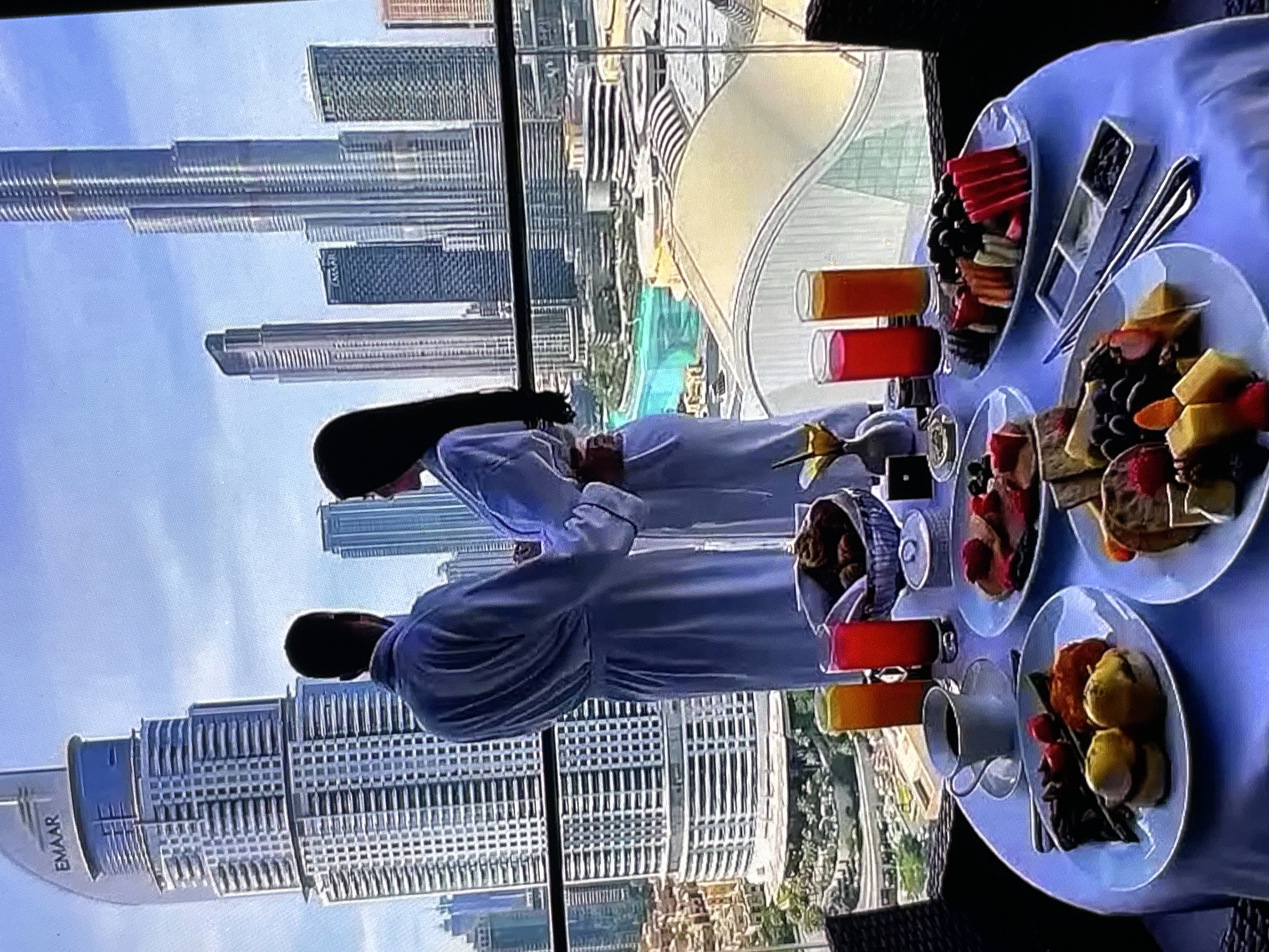 A couple stands on a balcony, both wearing white bathrobes. The male figure is seen from the back, with his left arm around the female figure. The female figure is viewed from the side, her head turned to her left. In the foreground, a table is set for breakfast. The table holds various dishes, including plates of food, a basket of bread, two glasses of juice, and a cup of coffee. The backdrop is a cityscape with numerous skyscrapers, characteristic of a large metropolitan area. The sky is clear and blue. The provided image does not contain any elements that would suggest the location is Yalga, Russia.