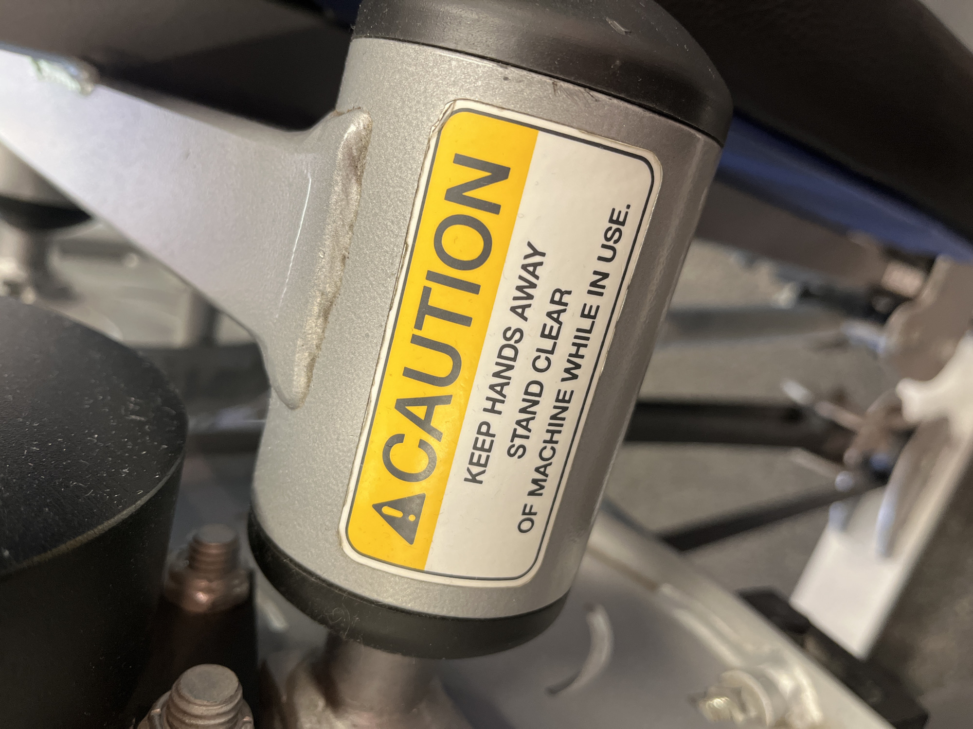 A close-up, eye-level shot focuses on a yellow and white caution sticker attached to a gray metal machine part. The sticker displays a black triangular warning symbol and the word "CAUTION" in large black letters. Below this, smaller black text reads "KEEP HANDS AWAY" on one line, and "STAND CLEAR OF MACHINE WHILE IN USE." on the next. The sticker is slightly wrinkled. The surrounding machine parts are metallic gray and black. The background is blurred, showing hints of other machine components and a dark blue surface. The scene is indoors, likely in a gym or workshop. No people are visible. The location in Saint Petersburg, Russia is not determinable from the image.