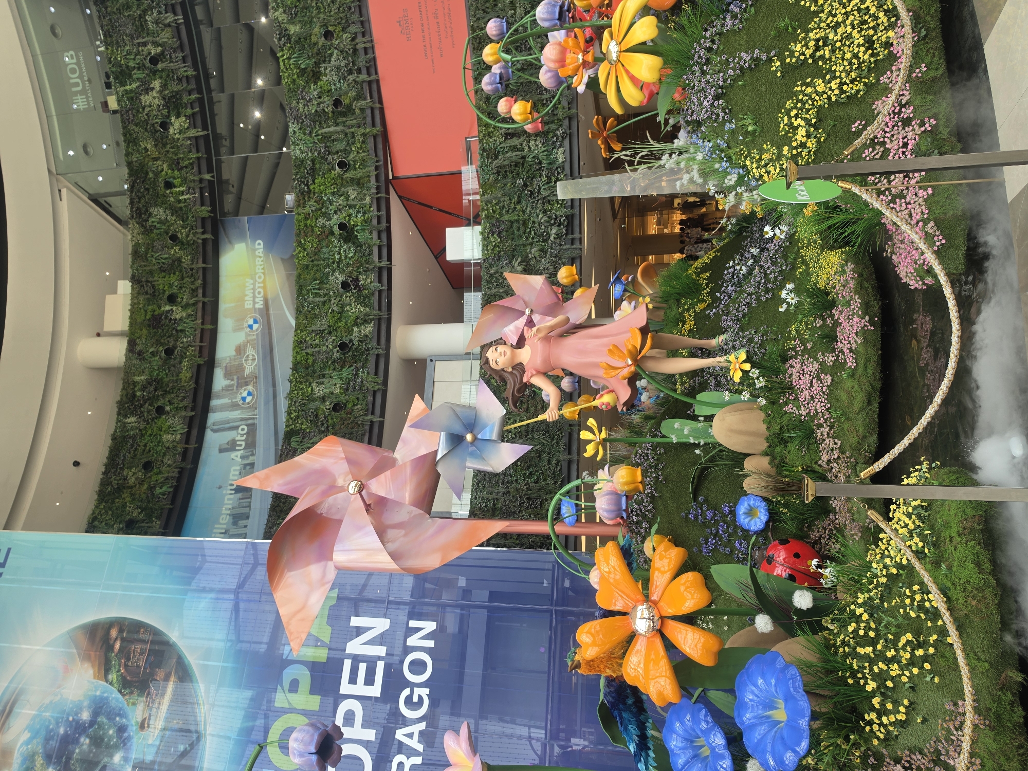 An eye-level shot captures a vibrant, whimsical indoor art installation, likely situated within a shopping mall in Bangkok, Thailand. The central focus is a large, colorful garden-themed display featuring a female mannequin or statue, resembling a fairy or nature spirit, dressed in a pink dress. She stands amidst giant, stylized flowers in shades of orange, yellow, and blue, along with large pink and blue pinwheel sculptures. A prominent red ladybug sculpture is also visible among the foliage. The ground of the display is covered in artificial green grass and dotted with numerous small, artificial flowers in yellow, pink, purple, and white, creating a lush, fantastical landscape. Mist effects emanate from the right side of the display, adding to the ethereal atmosphere. A braided rope barrier delineates the perimeter of the installation.
In the background, a multi-story building interior is visible. A large vertical garden or 'living wall' covers a significant portion of the background wall, contrasting with modern glass and metal architecture. Several large banners are present, displaying text such as "UOB WEALTH BANKING," "BMW MOTORRAD," and "Millennium Auto." Another banner shows "HERMES UNVEIL THE NEW CHAPTER" in English, followed by Thai text that translates to "Find Hermes store on floor." Partially visible text on a blue banner in the foreground reads "OPEN DRAGON" and "OP." The scene is brightly lit, characteristic of an indoor commercial environment, and conveys a cheerful and inviting mood. Blurry figures of people can be seen in the distance, observing the display.