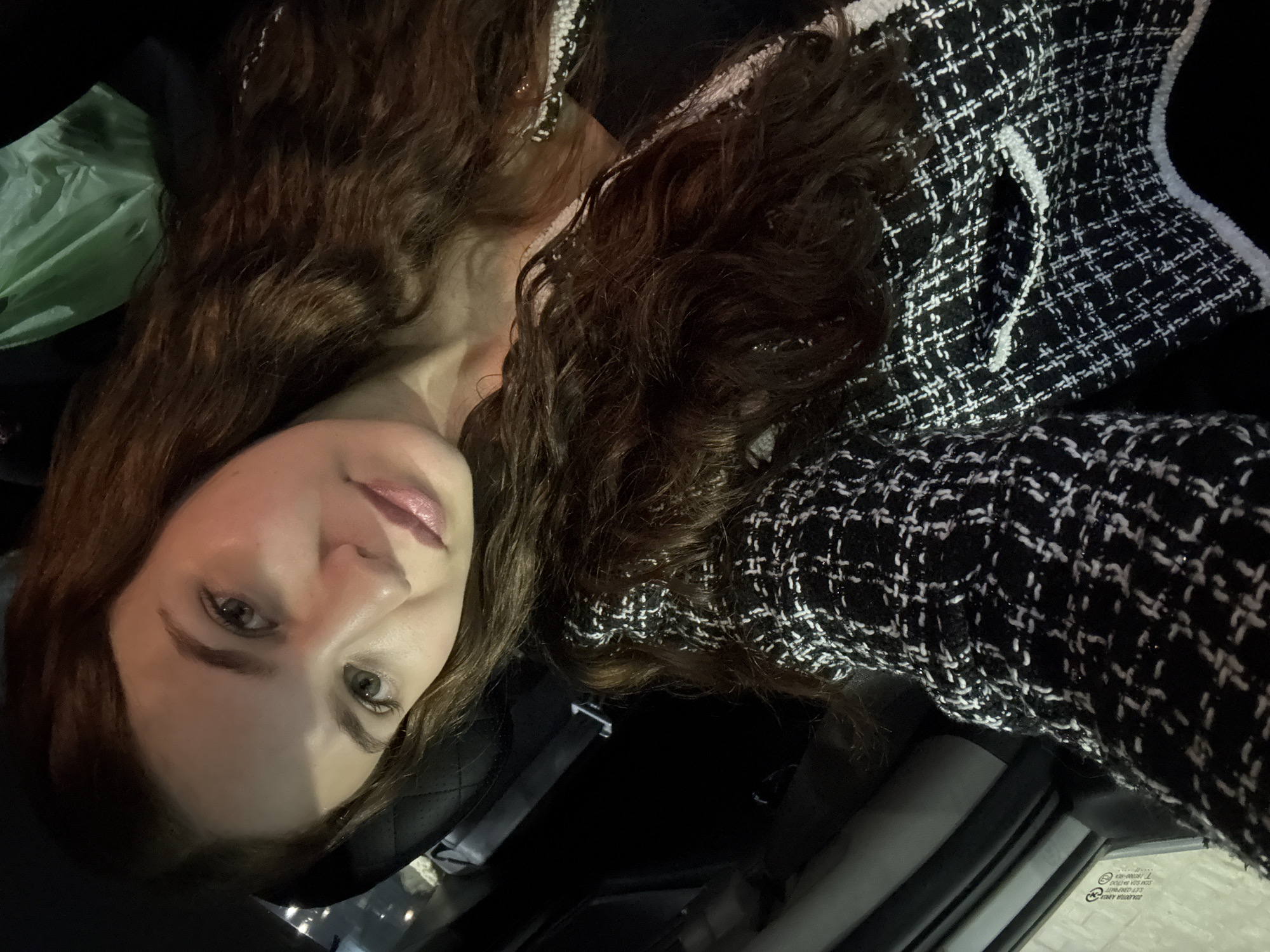 A woman with long, wavy brown hair, wearing a black and white tweed jacket and a black top, is pictured in what appears to be the interior of a vehicle. Her face is turned towards the camera, with her head tilted and resting on her shoulder. Her eyes are open and looking towards the viewer. The background is dark, with a glimpse of a green plastic bag visible behind her. The image is taken from a high angle, looking down at the woman. There are no discernible elements in the image that would indicate the location in the city of Chaikovsky, Russia.
