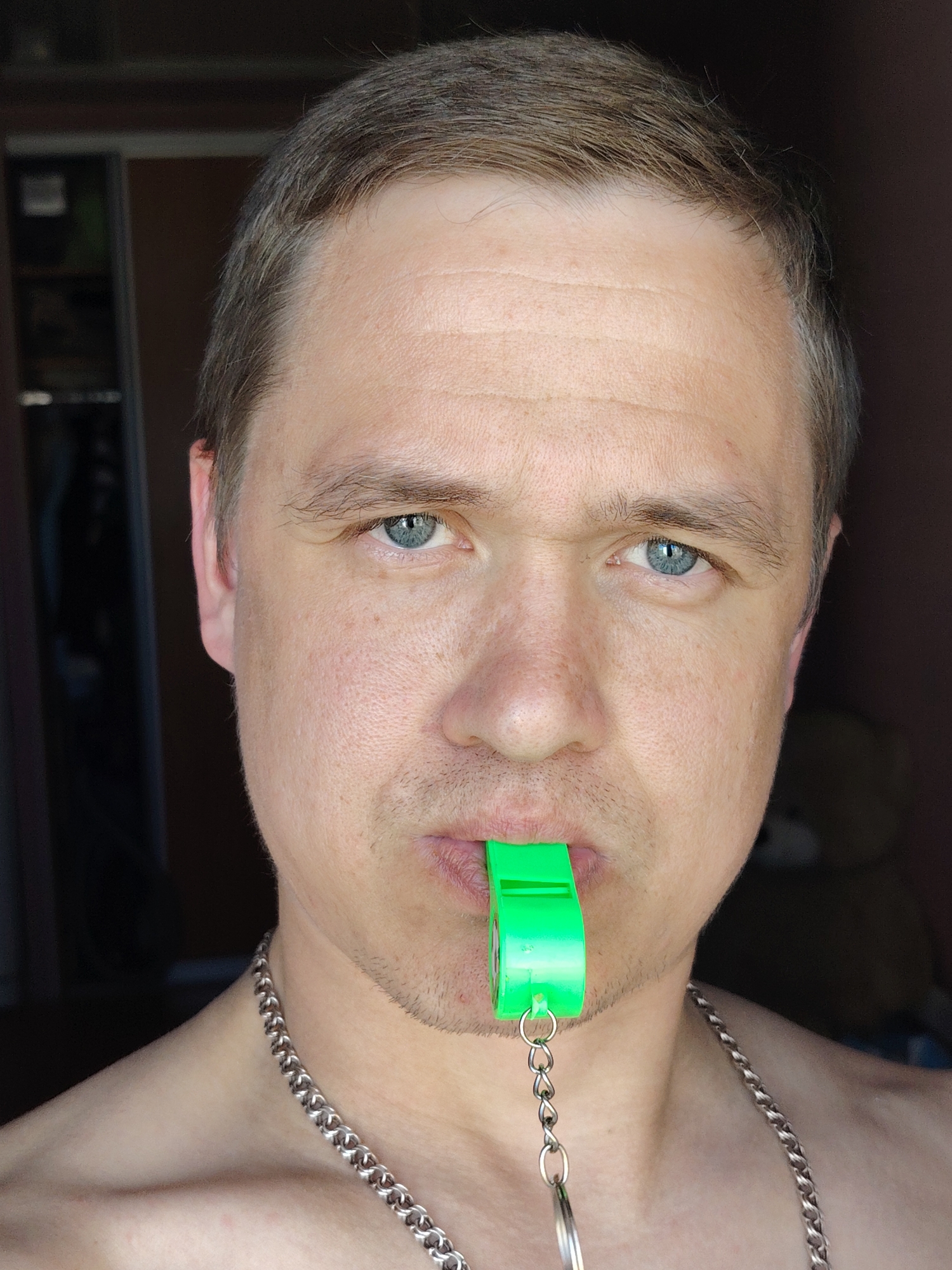 A close-up, eye-level shot features a Caucasian male with short brown hair and blue eyes, holding a green plastic whistle in his mouth. He wears a silver chain around his neck. The background includes a dark cabinet with a glimpse of items inside on the left, and a partially visible teddy bear on the right, against a dark brown wall. The image was taken indoors in a location that appears to be his residence. The city of Chaikovsky, Russia, is the potential location of the photo, but it does not appear in the image itself.