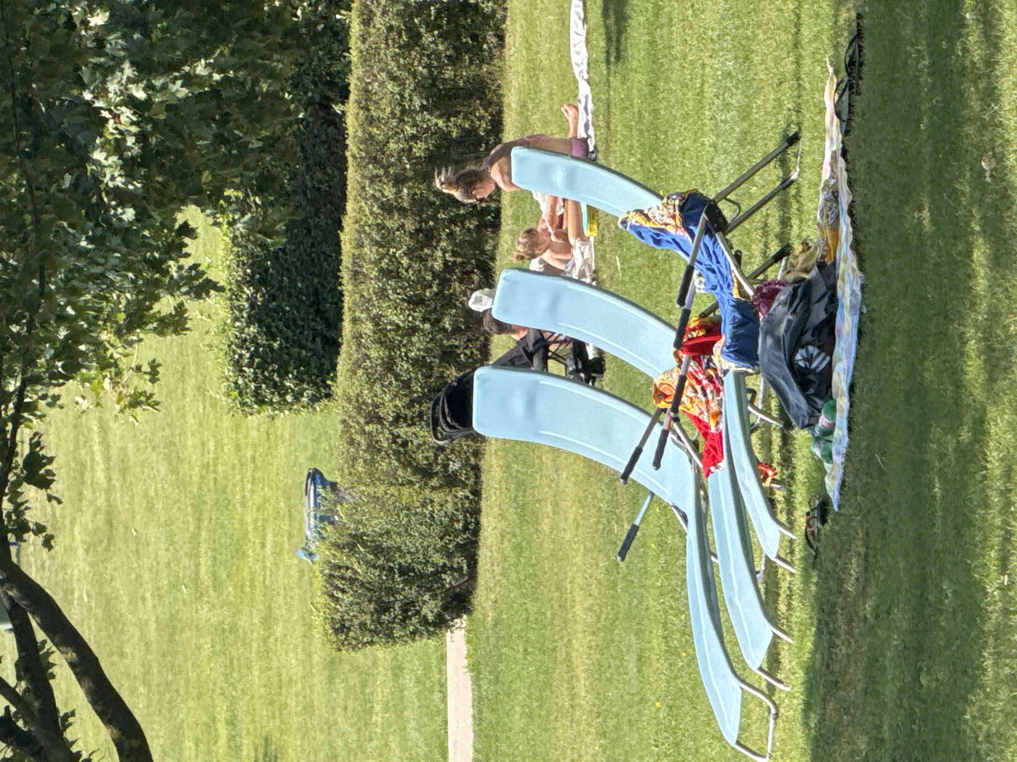 The image shows a sunny outdoor scene with several people relaxing on the grass. In the foreground, there are three light blue lounge chairs. Colorful towels and clothing are draped over the chairs and a nearby metal frame, suggesting they are drying or have been recently used. Two individuals are visible in the background, both appearing to be women. One woman is lying on a towel in the grass, wearing a bikini, while another woman is sitting in one of the lounge chairs, looking towards the ground. The setting is a well-maintained lawn bordered by a dense, manicured hedge. A tree with green leaves partially frames the left side of the image. The presence of lounge chairs and the sunny weather indicate a leisurely day, possibly at a park, resort, or a private garden. The lighting suggests it is daytime, likely late morning or afternoon, with clear skies. No text is visible in the image. The overall impression is one of relaxation and enjoyment of a warm day.