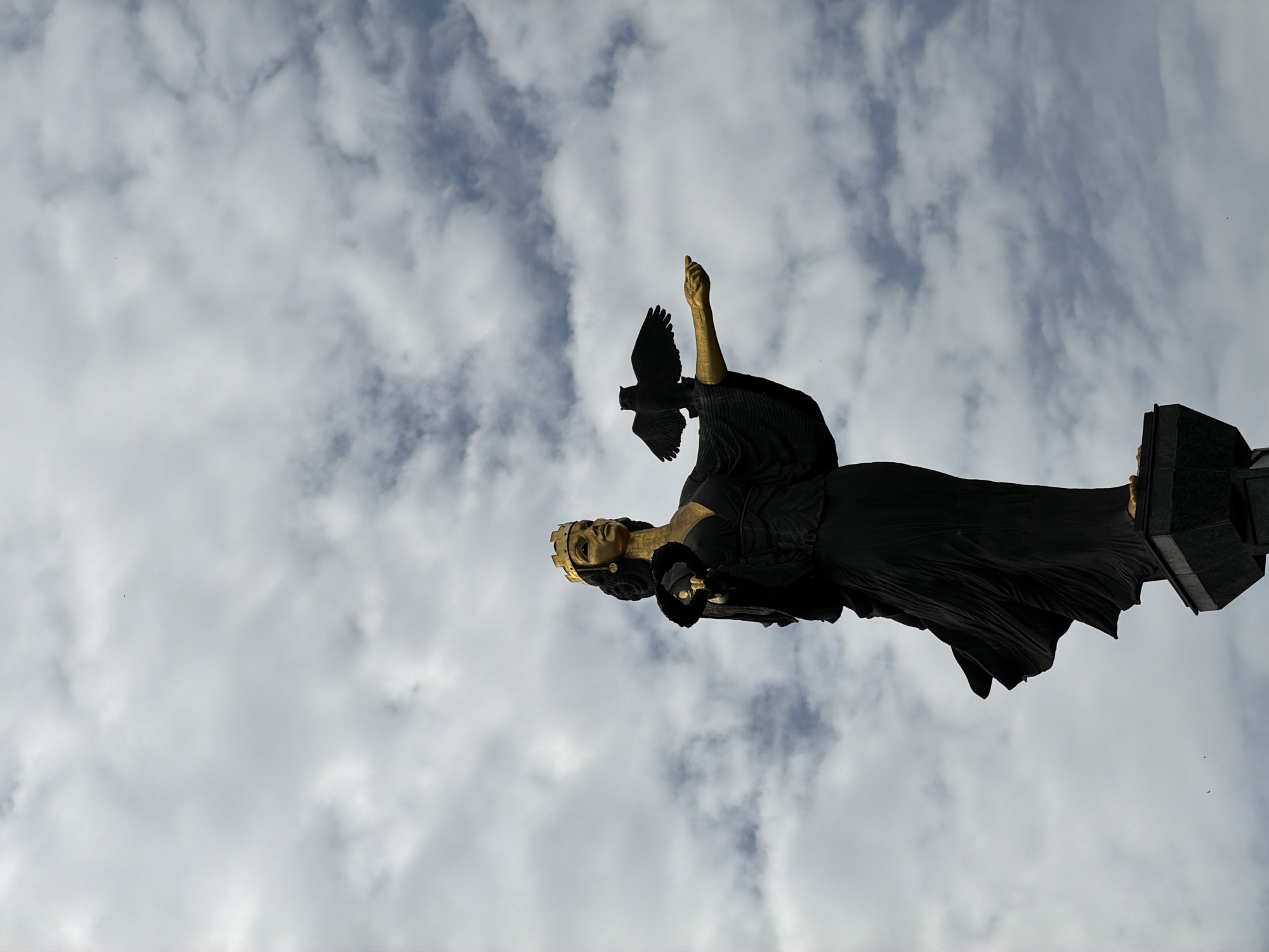 The image depicts the Monument of Saint Sofia in Sofia, Bulgaria, from a low-angle perspective against a cloudy sky. The monument is a dark, draped figure with golden hands, face, and crown. One hand is raised upwards, while a dark bird is positioned near the raised hand. The monument stands atop a dark, rectangular base. Small, dark birds are visible in the lower right corner of the sky.