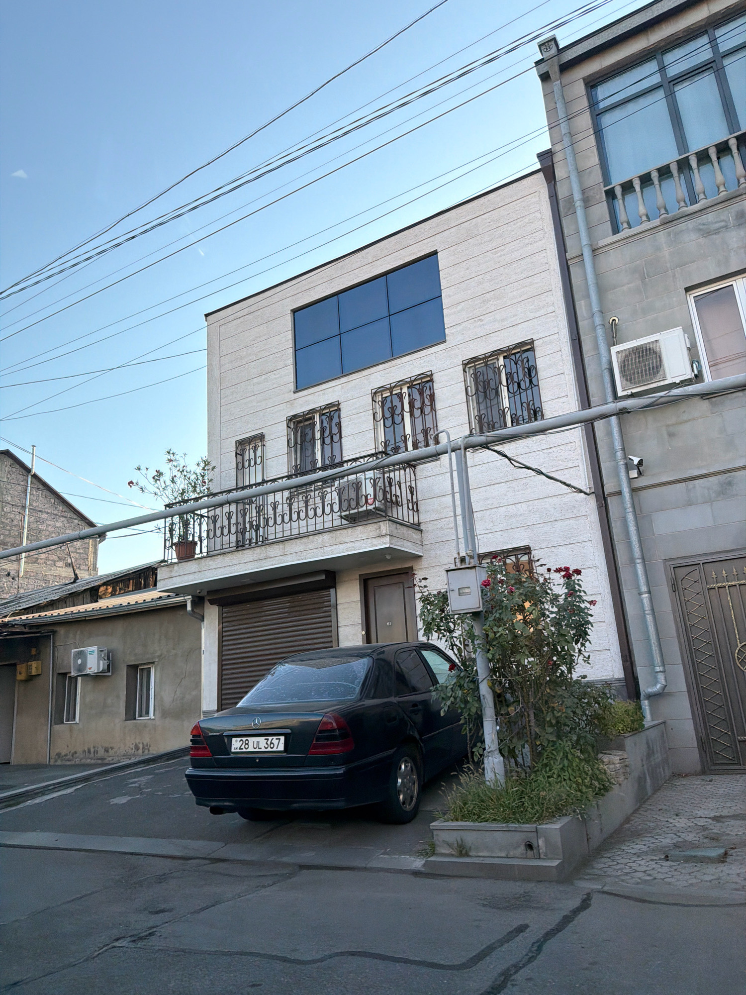 The image shows a street scene in Yerevan, Armenia, with a dark-colored Mercedes-Benz sedan parked on a paved surface. The car has a license plate reading "28 UL 367". To the right of the car is a modern building with a light-colored facade, featuring a large, dark, multi-pane window on the upper floor and a balcony with ornate metal railings on the middle floor. Below the balcony, there is a closed garage door. Adjacent to this building is another building with a grey, stone-like facade, featuring a balcony with white balusters and a window. An outdoor air conditioning unit is mounted on the grey building. In front of the modern building, there is a small planter box with a rose bush. Overhead, power lines are visible against a clear blue sky. To the left of the main building, there is a lower, older-looking structure with a grey concrete facade, a window, and another outdoor air conditioning unit. The overall environment appears to be a residential or mixed-use urban area.