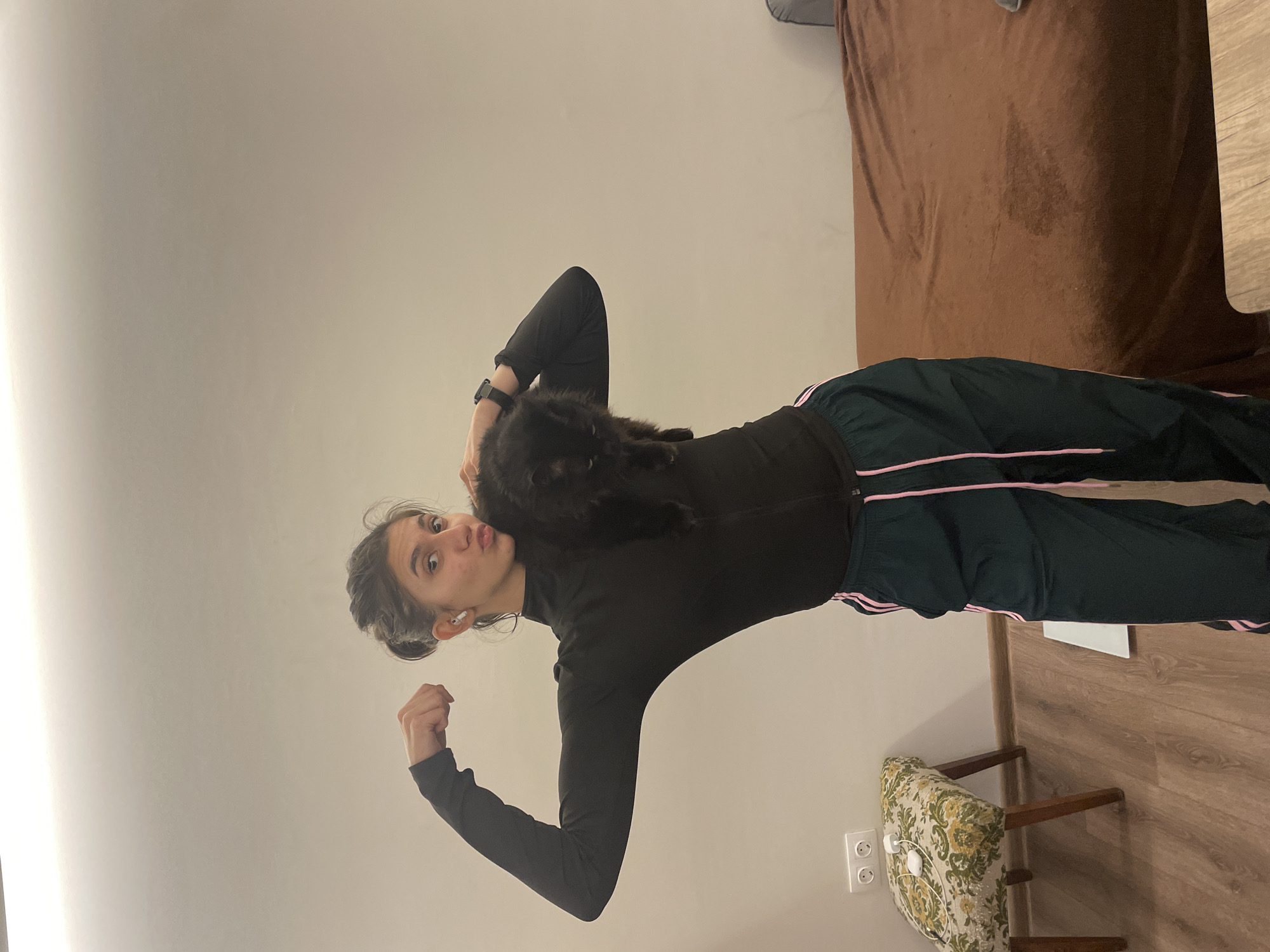 A woman is holding a black cat while posing in an interior setting. She is wearing a black top and dark green athletic pants with pink stripes. She is standing on a wooden floor next to a floral-patterned chair and a white electrical outlet on the wall. A brown bed is visible in the background. The location cannot be determined as Sofia, Bulgaria, based on the provided image.