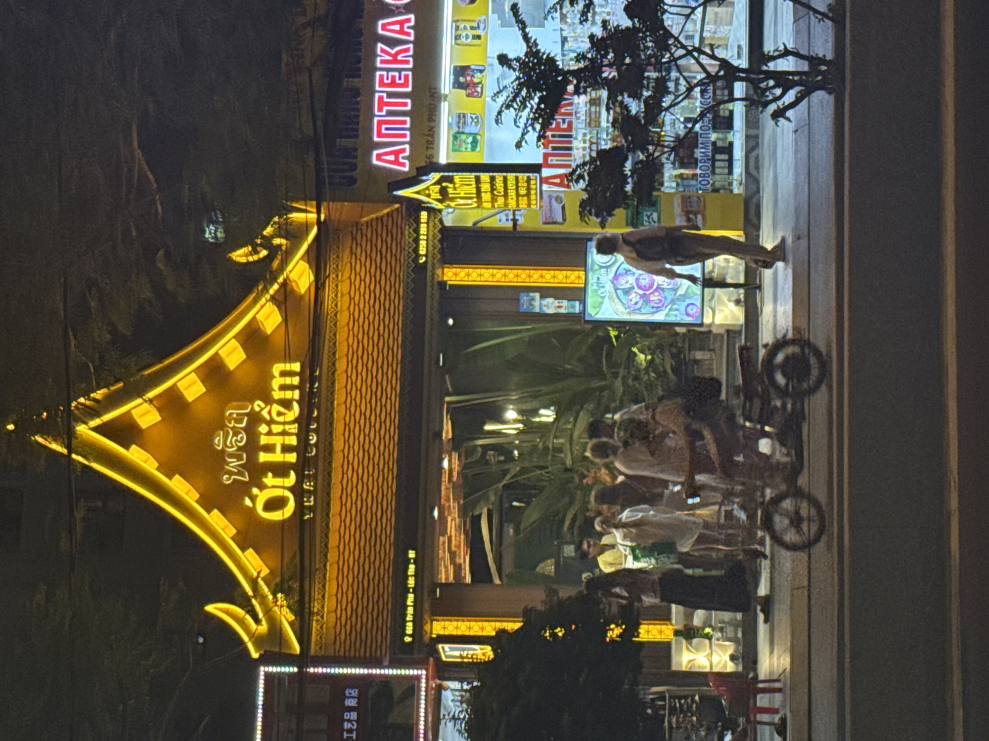 A nighttime street scene in Nha Trang Ward, Vietnam shows a group of people congregating on the sidewalk in front of the "Ớt Hiểm" restaurant. The restaurant's illuminated sign is a prominent feature, with golden lettering and ornate design. To the left of the restaurant is a sign indicating "9 GEA Trần Phú - Lớc Tho - BT" and another sign for "工艺品商店" (Crafts Shop). A lit sign for "АПТЕКА" (Pharmacy) is visible, with the address "66 TRẦN PHÚ-NT" and the phrase "Thai Cuisine ДАЙСКАЯ КУХНЯ" (Thai Cuisine Russian Kitchen). Another sign in cyrillic reads "ГОВОРИМ ПОД УС" (We speak under us). A man walks along the sidewalk. A parked bicycle is on the sidewalk.