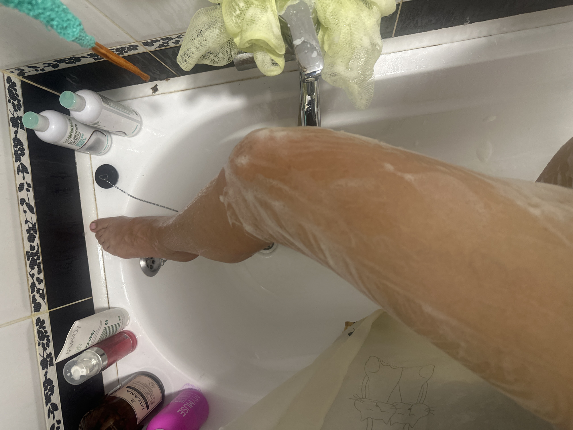 An overhead shot shows a person's soapy leg in a white bathtub. The leg is covered in white foam, and the person's foot is visible at the top of the frame, toes pointed upwards. Several toiletries are arranged on the edge of the tub, including bottles of shampoo and conditioner, a pink lotion bottle, and a brown bottle with a label that says "MILANA". A shower curtain with a cartoonish animal print hangs on the left side of the tub. A drain with a chain is visible in the center of the tub. A yellow loofah hangs from the faucet on the right side of the tub. The tiled wall behind the tub is black and white. The image was taken in Novoaltaysk, Russia.