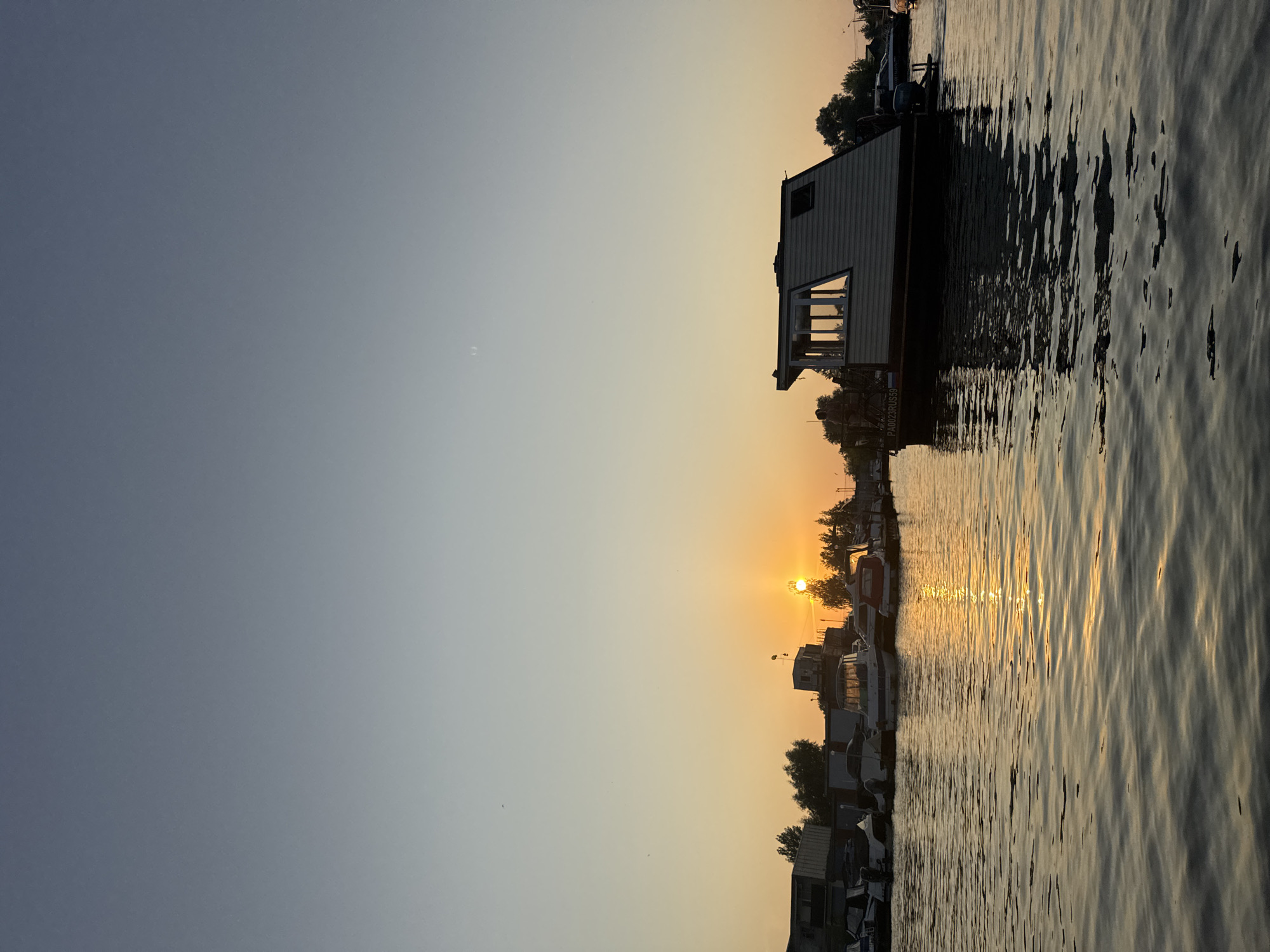 A wide shot captures a serene sunset over the Kama River in Chaikovsky, Russia, with the water reflecting the warm hues of the setting sun. In the foreground, the tranquil river flows, its surface rippling gently. Along the riverbank, a marina is visible, dotted with various boats. Notably, a distinctive house-like boat with a unique angular roof and large windows is moored in the middle distance, its silhouette sharp against the twilight sky. Several other smaller boats are docked alongside it. The background reveals a line of trees and buildings, contributing to the overall impression of a peaceful waterside settlement. The sky transitions from a pale blue at the top to a soft orange near the horizon, where the sun is setting. The scene is one of quiet repose, illuminated by the fading light of day.