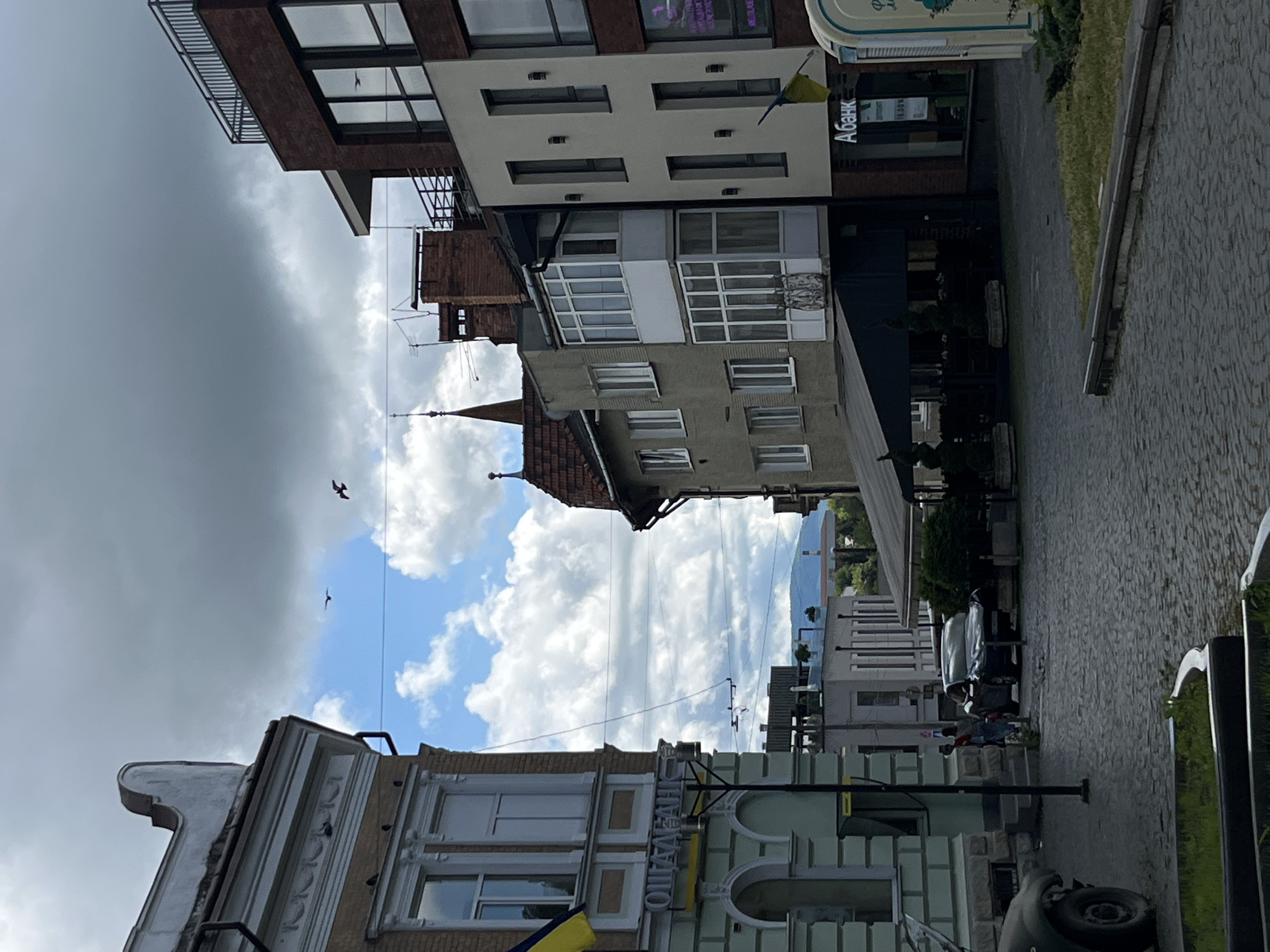 The image depicts a street scene in Drohobych, Ukraine, characterized by a mix of architectural styles. In the foreground, a cobblestone street leads towards several buildings. On the left, a light-colored building with a distinct architectural detail on its roof is partially visible. The word "ОЩАДБАНК" (meaning "Savings Bank" in Ukrainian) is visible on this building's facade. To the right, a modern brick building with large windows stands next to older, lighter-colored buildings featuring balconies and prominent chimneys. The sky is partly cloudy, with dramatic formations of grey and white clouds. Two birds are captured in flight against the sky. In the midground, a dark-colored car is parked on the street, with two people standing nearby. An awning covers an outdoor seating area in front of one of the buildings. A Ukrainian flag is visible on the modern brick building. The overall impression is of an urban environment with a blend of historical and contemporary structures under a dynamic sky.