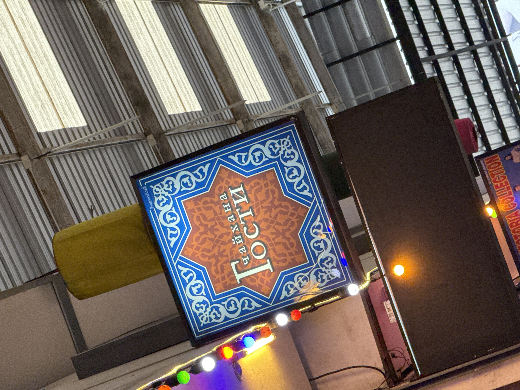 The image captures a sign for "Чайхана Гости" (Chaihana Gosty, meaning "Guest Tea House") hanging from a ceiling structure, likely in Karon, Thailand. The sign is rectangular with ornate blue and white patterns around a central octagonal design. The text "Чайхана Гости" is prominently displayed in white on an orange background within the octagon. Below this sign, a dark brown rectangular panel is visible, with a bright orange light emanating from its surface. String lights with various colored bulbs (red, green, blue, white, yellow) are strung to the left of the "Чайхана Гости" sign, illuminating the area. The ceiling consists of corrugated metal roofing with exposed beams and what appear to be skylights allowing natural light to filter through. A large, yellow cylindrical object is positioned above and to the left of the sign. The overall impression is of an indoor marketplace or restaurant setting.