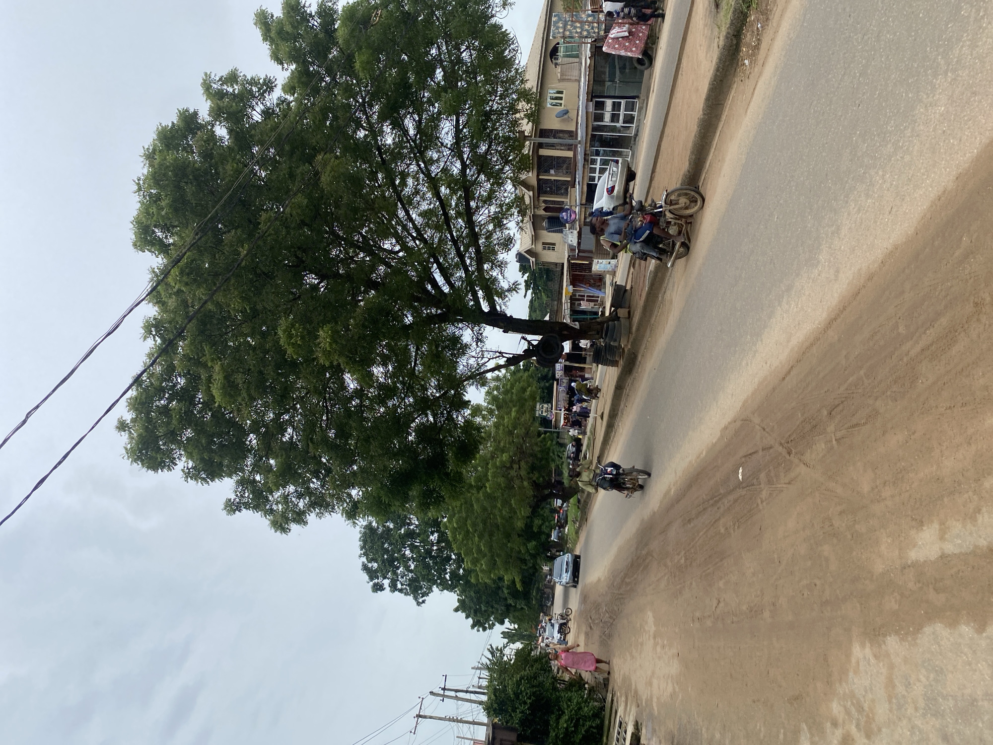 The image depicts a street scene in what appears to be a commercial or residential area. A large tree dominates the left side of the frame. Electrical wires run across the top left of the image. On the right side of the frame, there's a row of buildings, with shops and residential units. A motorcycle with passengers is parked along the curb. Other motorcycles and a car are visible further down the street. A person in a pink dress is walking on the right side of the street. The road itself appears paved, although partially covered in dirt. Based on the style of the buildings, vegetation and general atmosphere, the location might be Lagos, Nigeria.