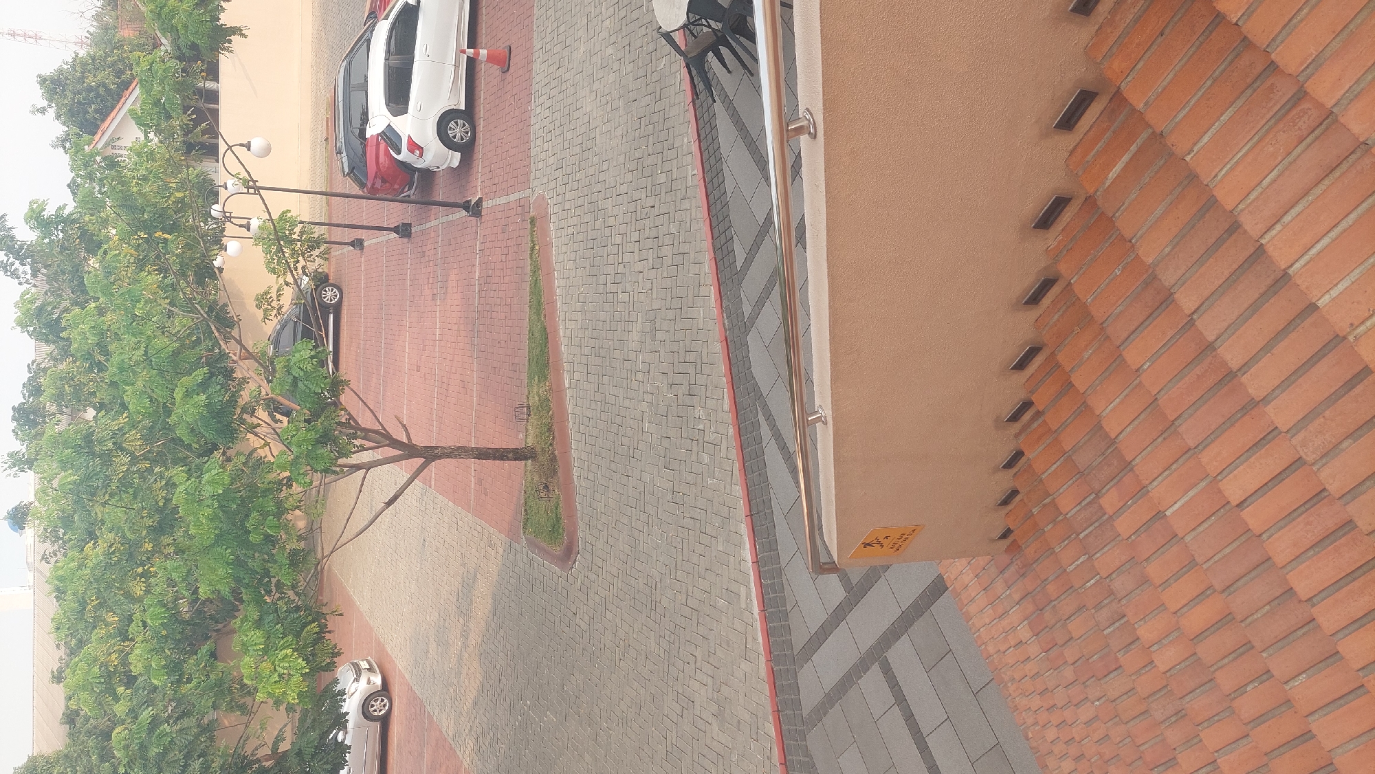 The image shows a view from a brick staircase looking down into a parking lot and a tree-lined area. In the parking lot, there are several cars parked, including a silver sedan, a black SUV, a red SUV, and a white hatchback. A bright orange traffic cone is visible near the white hatchback. Multiple street lamps with round white globes line the edge of the parking lot. A tree with green foliage stands in the middle of the parking lot. The parking lot is paved with red bricks and gray pavers. Adjacent to the parking lot is a sidewalk and a building with a light-colored wall. A small yellow sign with black text is attached to the wall near the top of the stairs. The staircase itself is made of reddish-brown bricks and has recessed lighting fixtures along the side wall. The environment appears to be an outdoor commercial or residential area in Cileungsi, Indonesia, likely during the daytime.