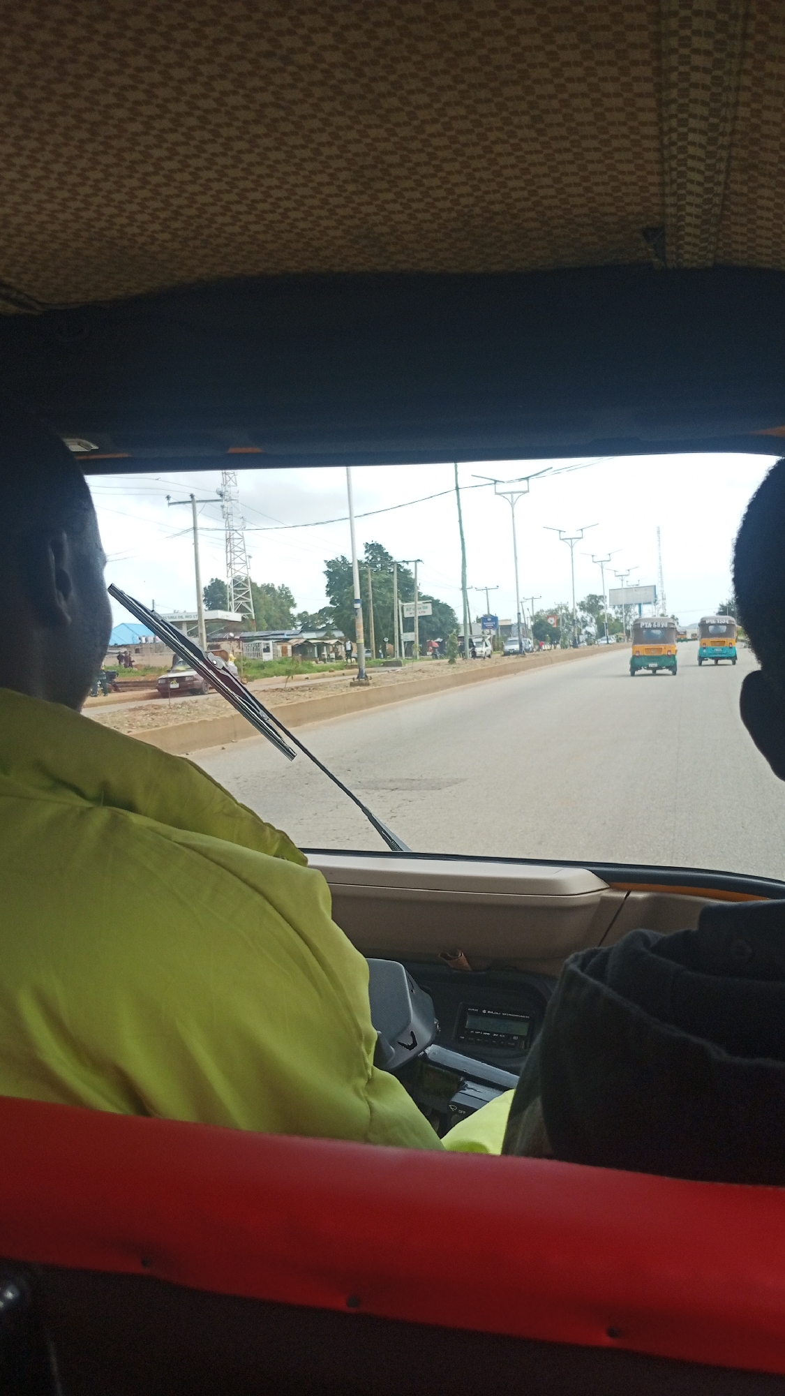 The image is taken from inside a vehicle looking out through the windshield. In the foreground, the back of the heads of two individuals is visible, partially obscured by the vehicle's interior. One individual in the driver's seat wears a bright yellow-green jacket. The windshield wipers are in the down position. The road ahead is paved, with a median strip separating lanes of traffic. On the left side of the road, there are small buildings, some appearing to be shops or residences, and a large telecommunications tower. On the right side of the road, there are streetlights and more buildings. Several vehicles are on the road, including two auto-rickshaws (Keke NAPEPs) in the distance. The overall environment suggests a busy urban or semi-urban area in Nigeria, consistent with Bukuru. The sky is overcast. The vehicle's interior shows a checkered patterned headliner and a dashboard with some electronic equipment. The scene depicts a view of a street in Bukuru, Nigeria, from the perspective of a passenger or driver in a vehicle.
