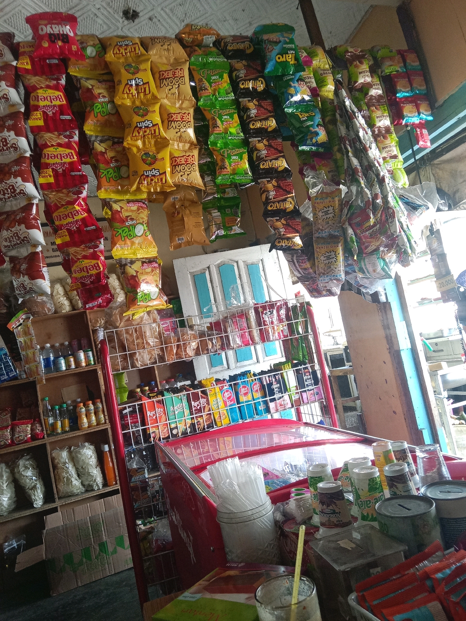 The image depicts a small, cluttered roadside convenience store in Kertek, Indonesia. Shelves are packed with a variety of snacks, drinks, and household items, including bags of chips, bottled beverages, and packaged goods. A red refrigerator case is partially visible, filled with more products. The overall impression is one of a busy, well-stocked shop with limited space. There are no people visible in the image, and the focus is entirely on the merchandise and the store's interior. The lighting is somewhat dim, suggesting it might be an overcast day or the store is located in a shaded area. The environment appears to be an indoor setting, possibly an open-fronted stall or a small room.