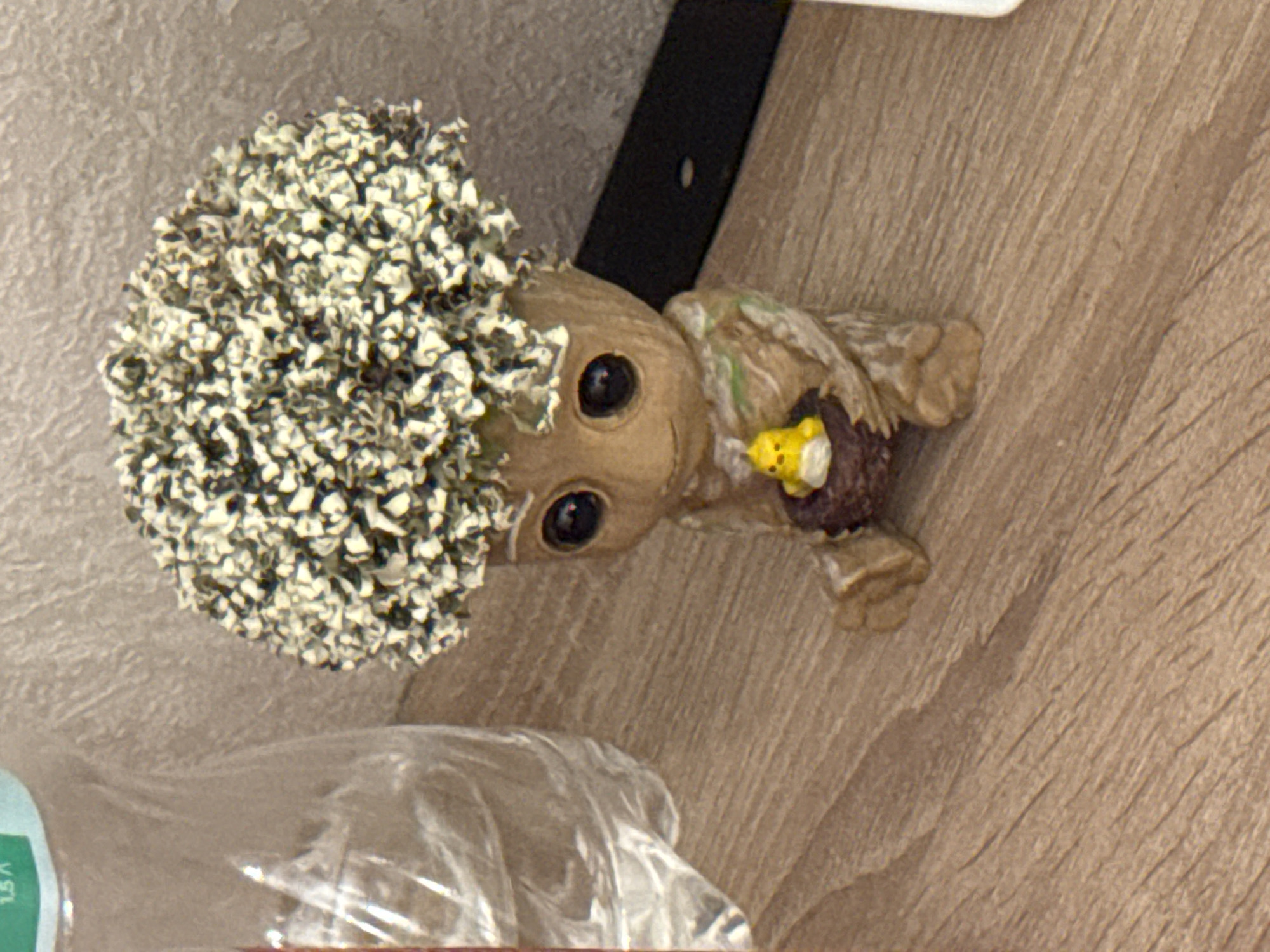 This is a close-up, eye-level, indoor shot of a decorative figurine resembling Groot from Guardians of the Galaxy, with a fluffy, textured green and white "hair" on its head. The figurine is made of a light brown material with a wood-grain texture and has large, dark eyes. It is sitting with its legs tucked beneath it and is holding a small, brown bird's nest in its lap. Inside the nest is a tiny yellow chick figurine. The figurine is placed on a wooden surface with a pronounced wood-grain pattern. To the left of the figurine is a portion of a clear plastic bottle, possibly containing water, with some writing visible. The background is a textured, light-colored surface, possibly a wall. There is no indication of the city of Saratov, Russia, or any other geographical location in the image.