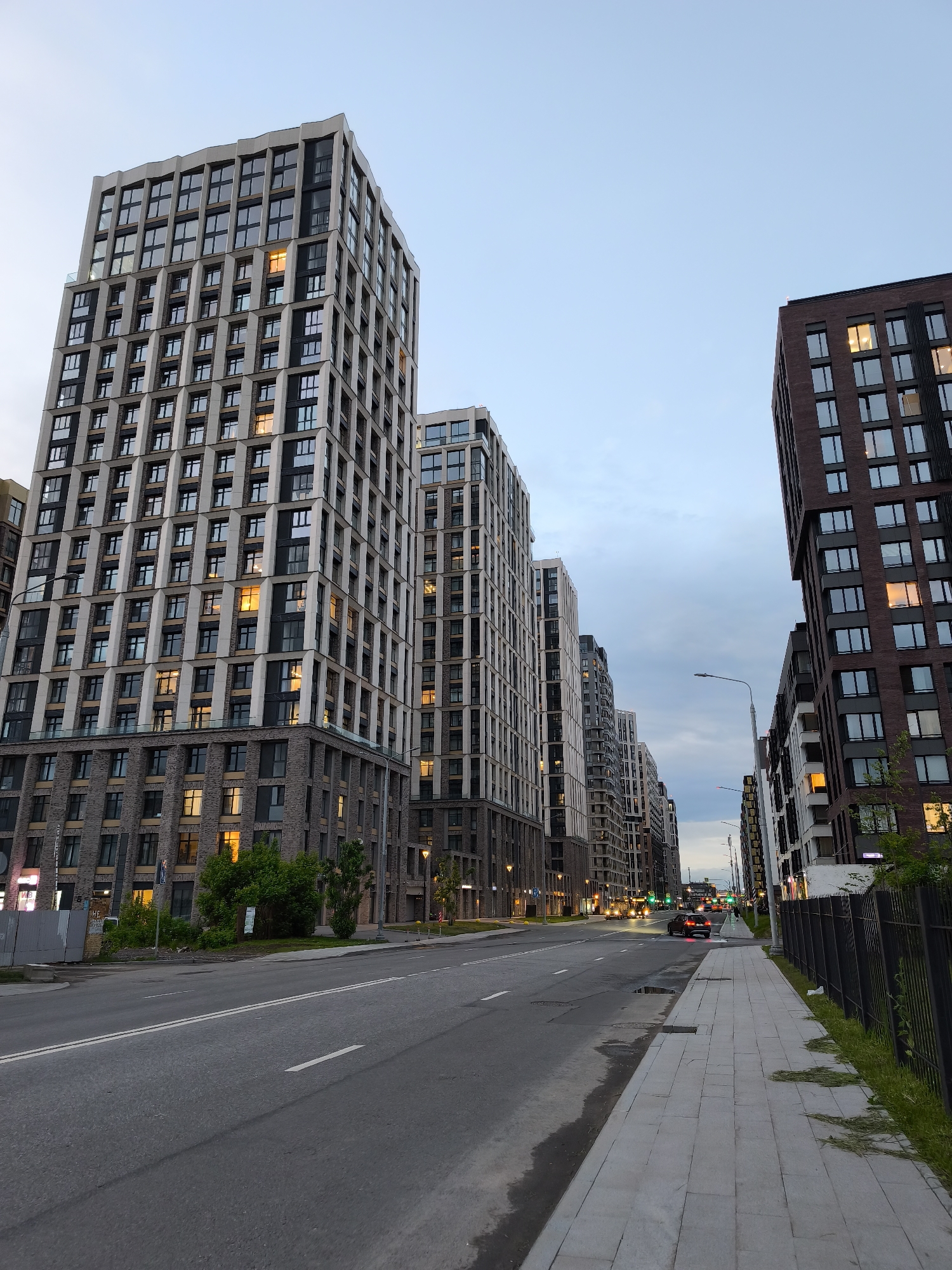 The image shows a street scene in Moscow, Russia, likely in a newer, upscale residential area. The street is wide, with a smooth asphalt surface and clearly marked lane dividers. A sidewalk runs along the right side of the street, bordered by a black metal fence.
Several modern, high-rise apartment buildings line both sides of the street. The buildings are constructed in a contemporary style, featuring repetitive window patterns and neutral-toned facades with dark accents. Some windows are illuminated, indicating occupancy.
Several cars are driving down the road, including a black SUV in the center lane and other vehicles further down the street. Streetlights are visible along the roadside. The sky is overcast, suggesting dusk or early evening. No pedestrians are visible on the sidewalk or in the street. The overall impression is of a quiet, modern urban environment.
