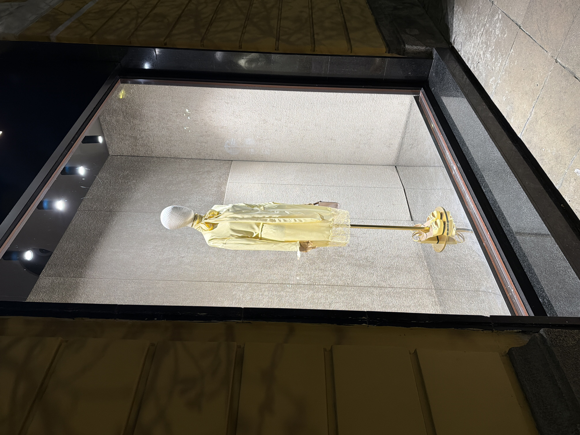 Captured from an oblique, elevated perspective, the scene reveals a window display in what appears to be an upscale retail setting. Inside the window, a mannequin is mounted horizontally on a gold-colored stand. The mannequin, draped in a light yellow garment, has a stark white head. The backdrop is a uniform, light-colored wall, likely constructed from stone or a similar material. A series of spotlights are attached to the interior of the window frame, casting light onto the display. The exterior of the window is characterized by a combination of dark stone and a lighter, textured material, which appears to be the facade of the building. The location within the city of Unknown, Unknown cannot be determined from this single image. The overall lighting suggests it is either dusk or night.