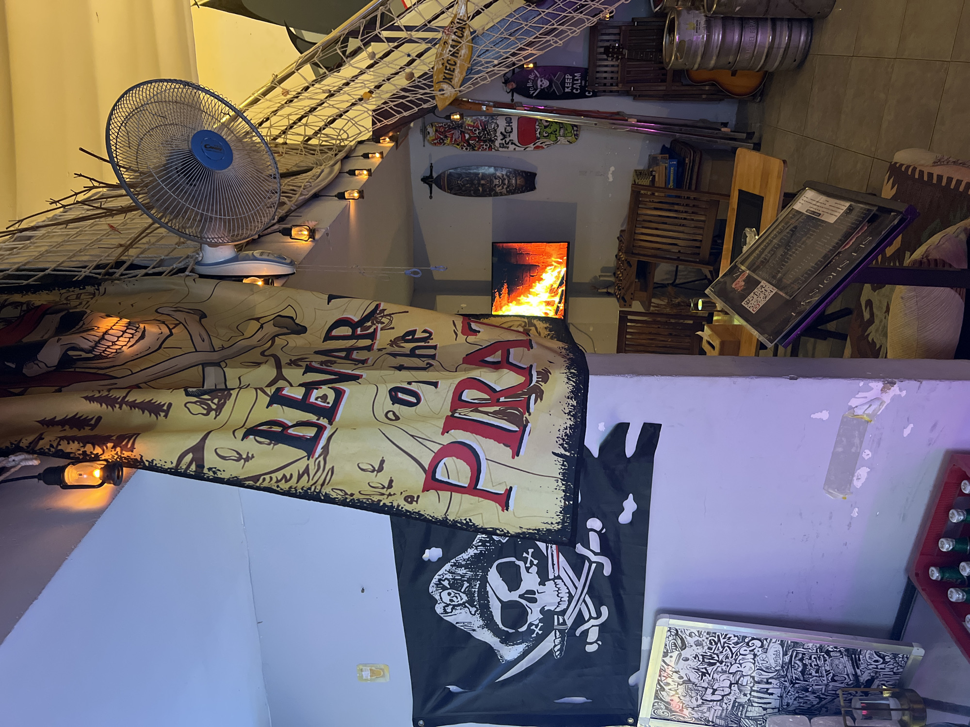 A low-angle indoor shot depicts a pirate-themed decor in what appears to be a restaurant or bar, with pirate flags and a "Beware of the Pirates" banner prominently displayed. A blue fan sits on a white ledge. The decor includes fishing nets, wooden planks, and string lights, creating an ambiance. Kegs and a guitar are visible in the background, along with skateboards and a TV screen showing flames. A menu holder stands on a table. A case of bottled drinks is also apparent. The location is likely Bắc Nha Trang, Vietnam, given the Vietnamese writing on one of the signs.