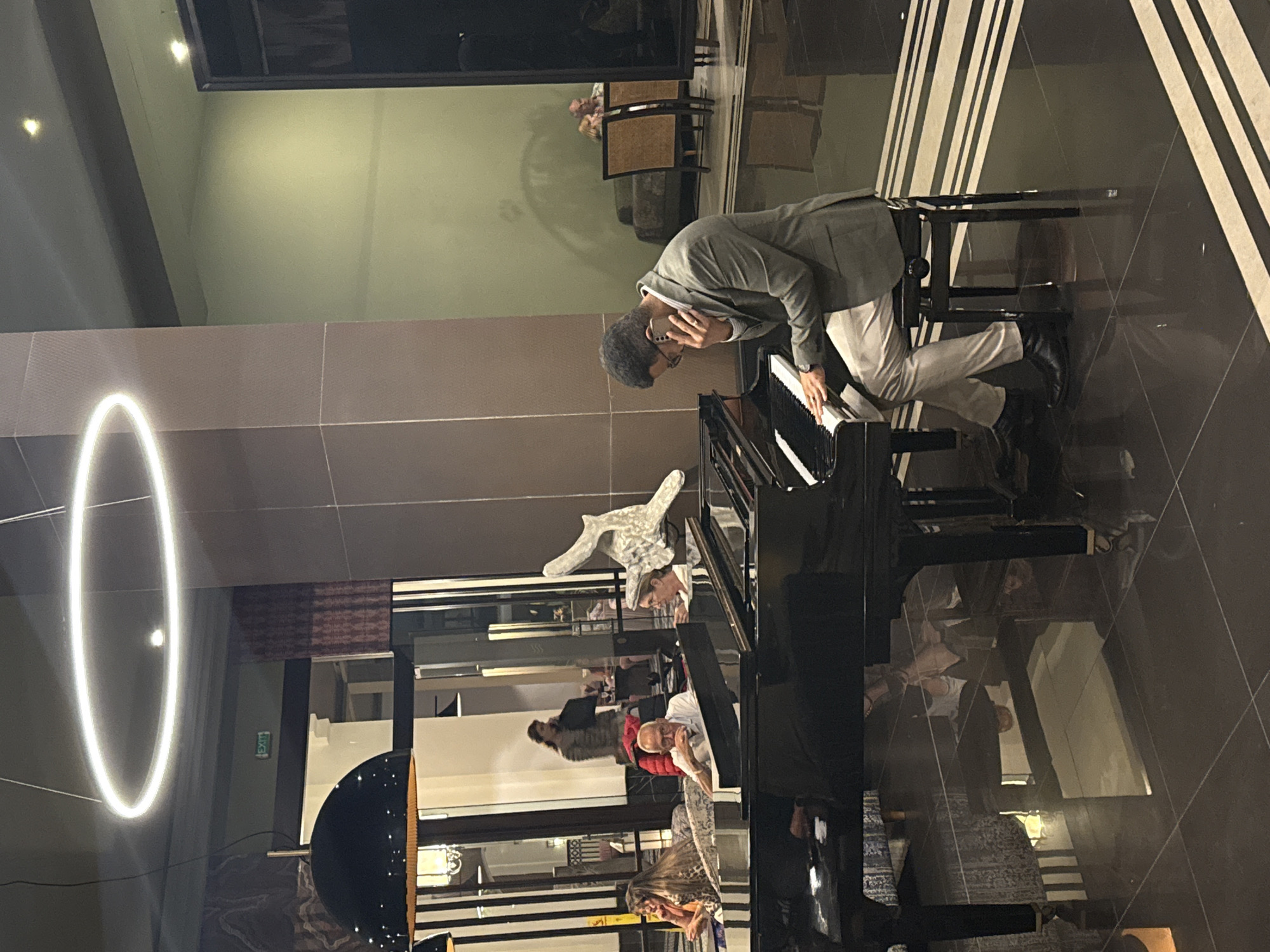 A medium, tilted shot captures a person in a gray suit playing a grand piano in what appears to be a hotel lobby. The person is leaning over the keyboard, seemingly focused on playing. Behind the piano, there are other people present, some sitting and some standing. The environment features modern décor with a large circular light fixture, reflective flooring, and striped designs on the walls. Given the upscale lobby setting and the person playing the piano, it's plausible the location is a luxury hotel in Agadir, Morocco, although specific landmarks are not visible to confirm the city.