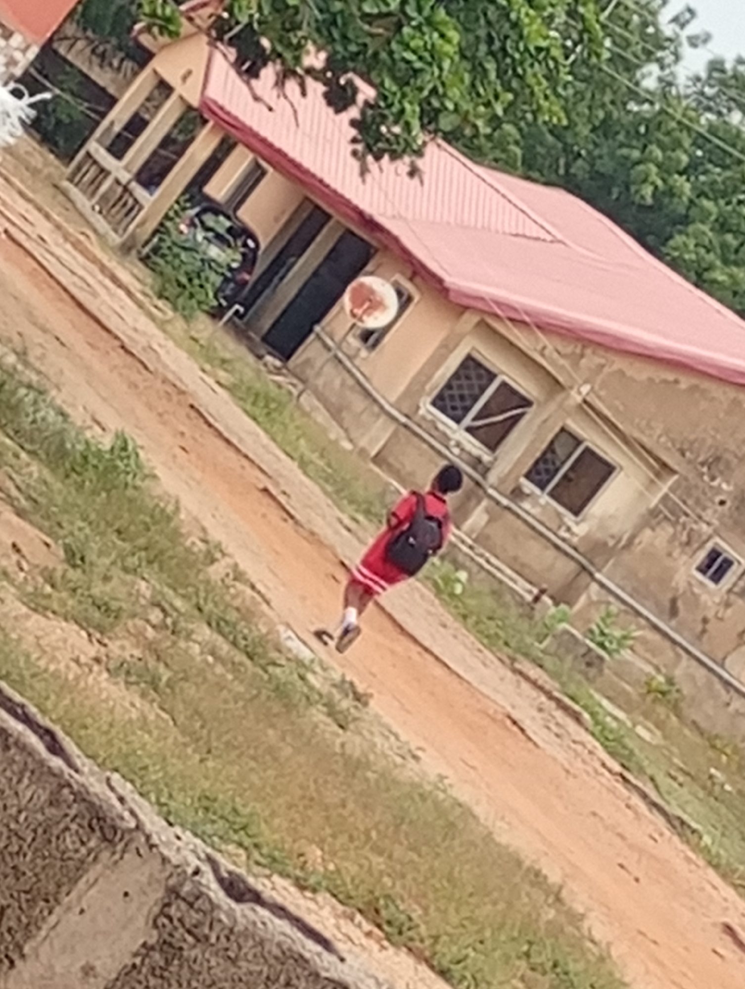 A person with a backpack walks on a dirt path near a house with a red roof, located in an unknown area within the city of Unknown, Unknown. The house has barred windows and a satellite dish. A car is parked in a partially visible garage. The environment includes vegetation along the path and near the house. The scene appears to be a residential area.