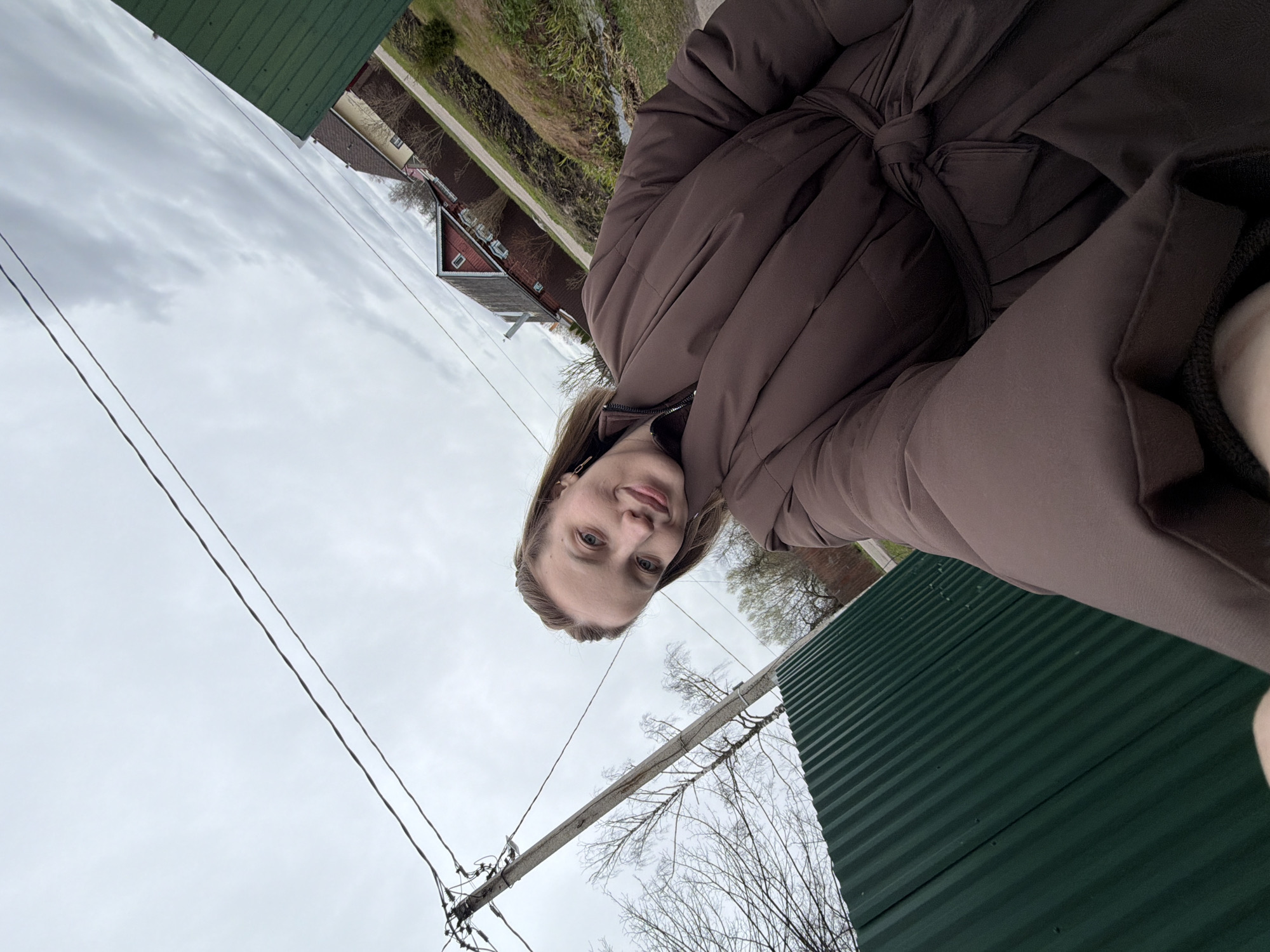 A person, likely female, is wearing a brown, quilted coat with a belt, taken at an acute angle upward. The background features a cloudy sky, overhead power lines, and glimpses of buildings and trees. At the right is a green corrugated metal fence. The environment suggests a suburban or rural area. The image does not contain enough information to determine whether it is in the city of Reshetnikovo, Russia.
