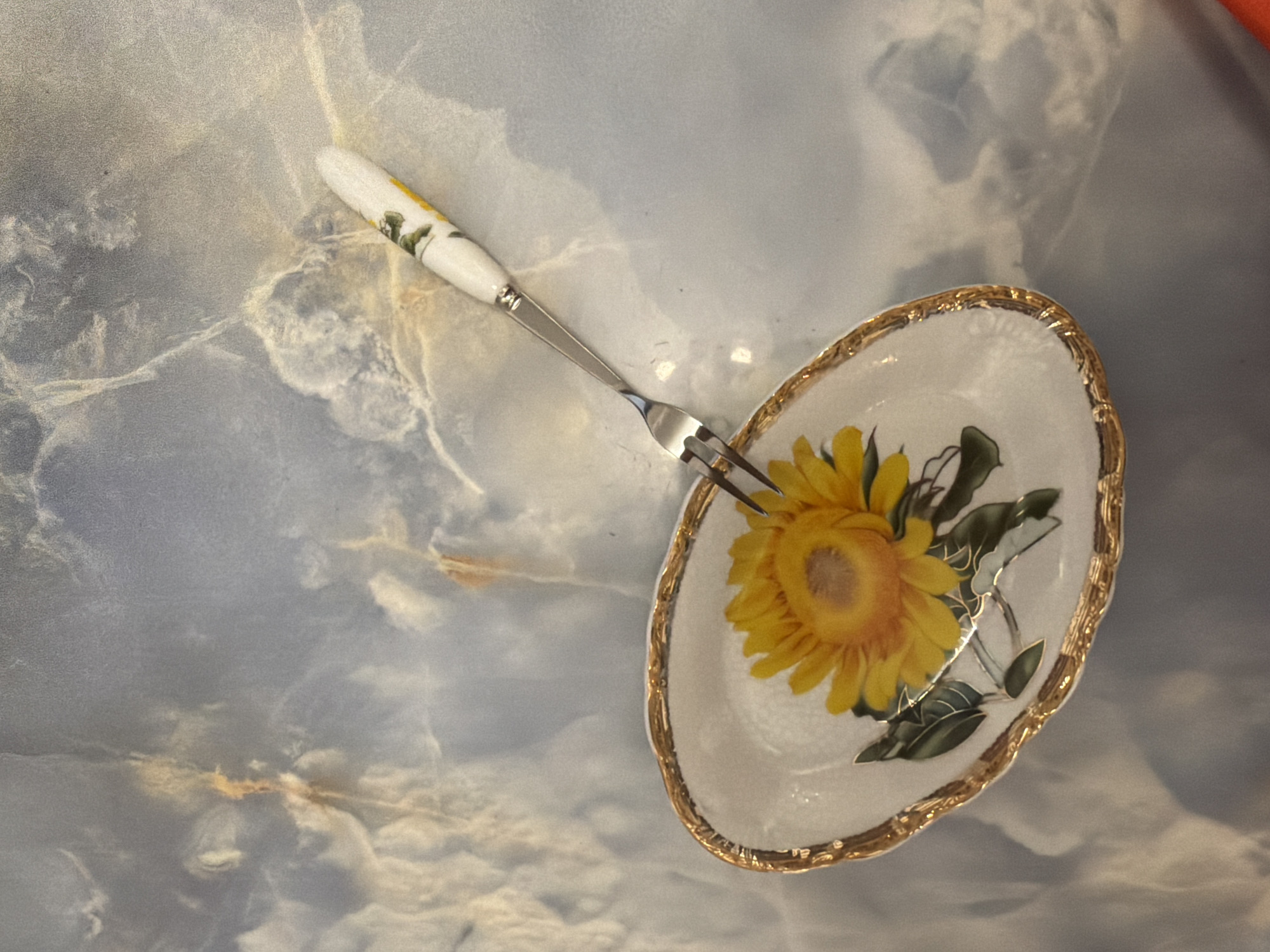 A fork with a white handle decorated with flowers rests on a white, oval dish with a gold rim, containing a painted sunflower with green leaves. The dish and fork are placed on a reflective surface with a blue and white marbled pattern.