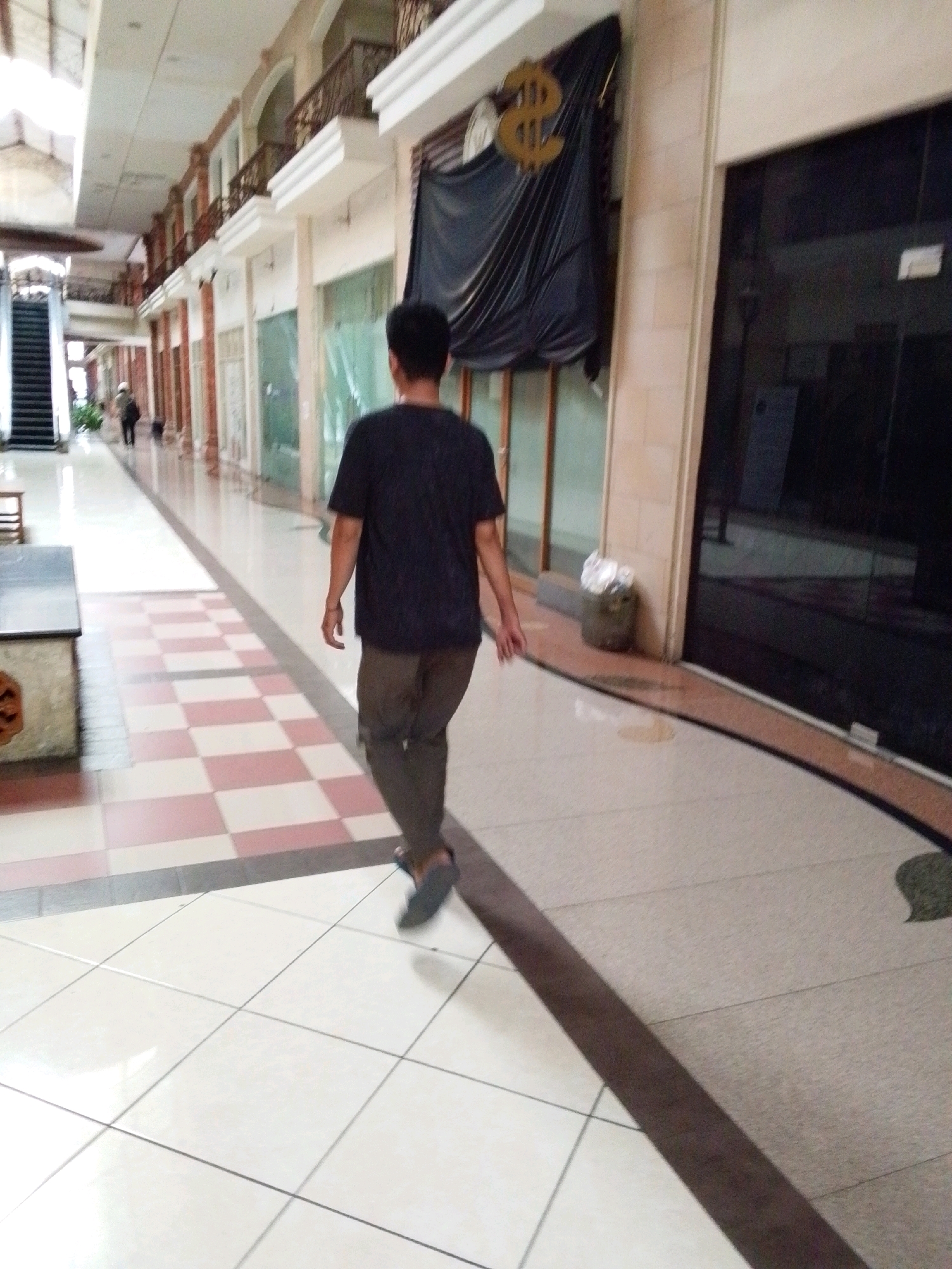 The image depicts the interior of a shopping mall in North Jakarta, Indonesia. A lone individual, viewed from behind, walks away from the camera down a wide, tiled corridor. The person is wearing a dark t-shirt and olive-colored pants. The corridor features a checkered floor pattern of light pink and white tiles, transitioning to larger white tiles closer to the viewer. On the left, an escalator is visible, and further down the hall, another person in dark clothing is seen walking. Shops with reflective glass storefronts line the right side of the corridor. A black tarp with a gold dollar sign is draped over a structural element on the right wall. The environment appears somewhat dated, with some visible wear and tear on the flooring.