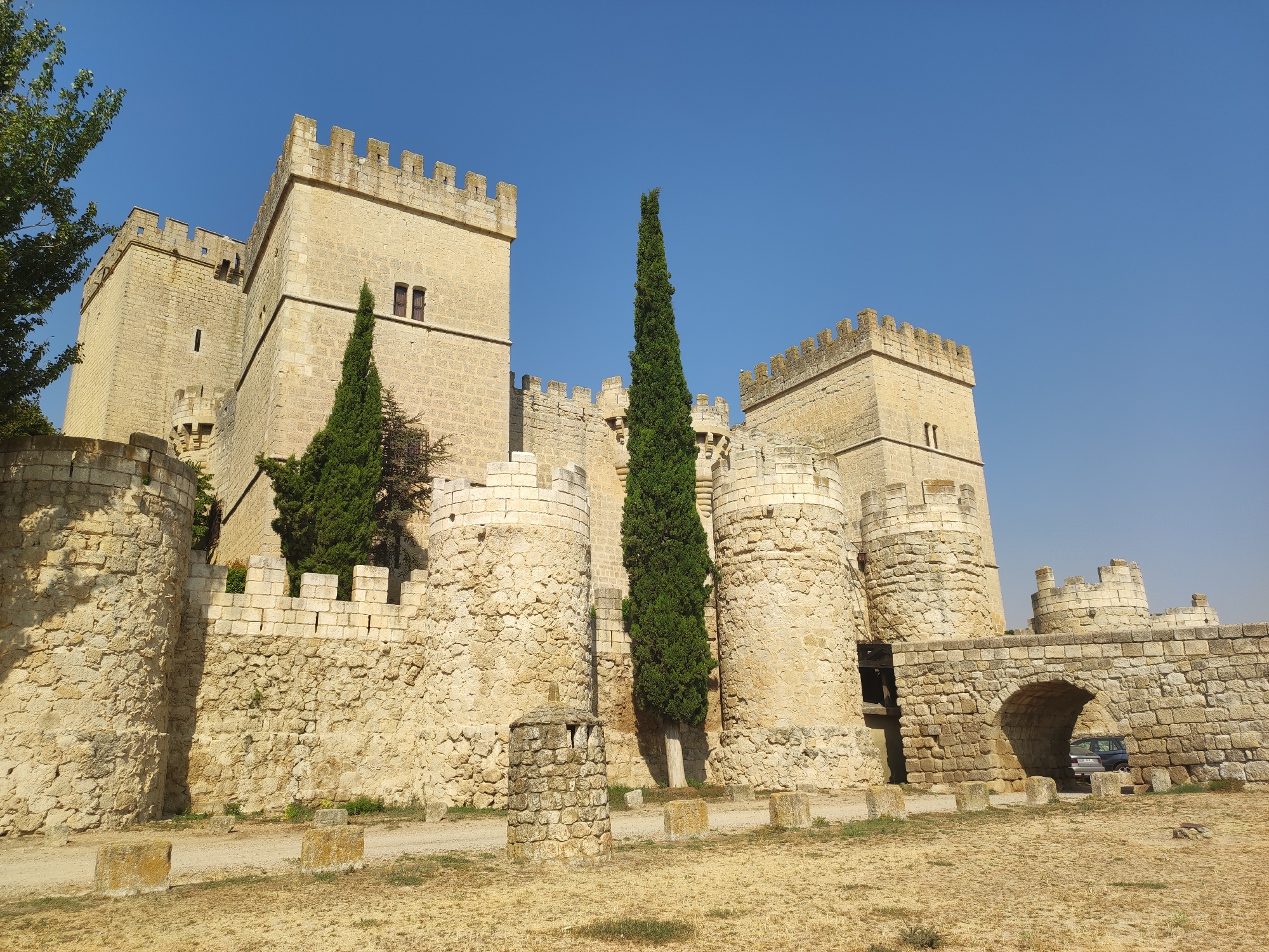 This is a medium shot of the exterior of the castle of Ampudia in Spain, under a clear blue sky. The castle is constructed of light-colored stone and features crenellated battlements atop its towers and walls. The main structure is a large, rectangular keep with a smaller tower attached to its side. In front of the keep, a section of the castle wall is visible, constructed of rough-hewn stones. Several round towers with crenellated tops are also part of the castle's defensive perimeter.
To the left of the keep, a large tree with green foliage partially obscures the view. Two tall, slender cypress trees stand in front of the castle walls, one centrally located and another to the left. The foreground consists of a dry, sandy ground with scattered stones and some patches of dry grass. A stone archway, part of the castle's outer defenses, is visible on the right side of the image, with a dark-colored car parked beyond it. The overall impression is of a well-preserved medieval fortress under a bright, sunny sky.