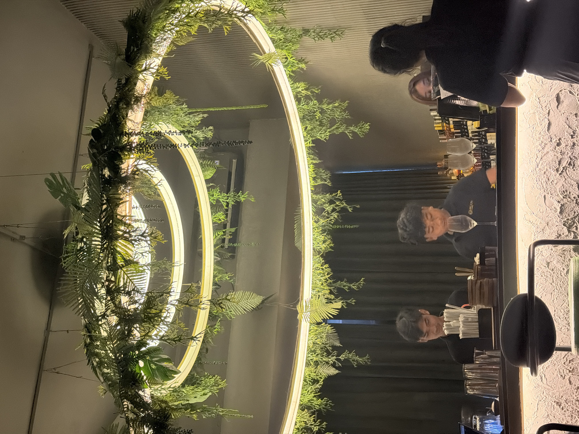 In North Kuta, Indonesia, a dimly lit bar scene unfolds beneath a large, circular light fixture adorned with artificial greenery. Two male bartenders, dressed in black shirts, are visible behind the counter, preparing drinks. One is holding a glass, while the other appears to be arranging straws. To their right, a female patron is seen looking down at something on the bar. Bottles of alcohol line the shelves behind the bar. The bar itself has a textured, illuminated front. A woman's back is visible in the foreground, facing away from the camera and towards the bar.