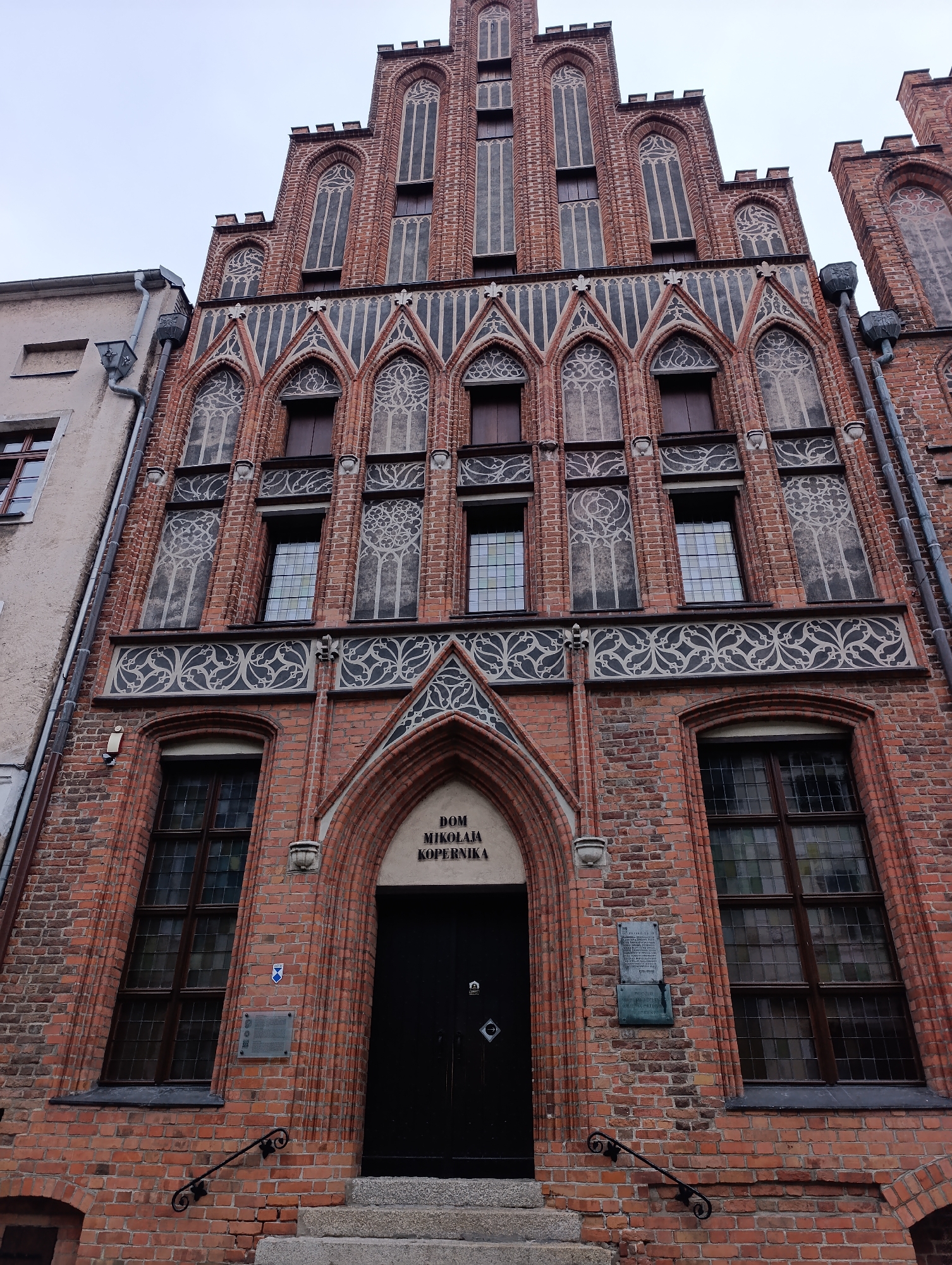 The image shows the facade of a historic brick building in Toruń, Poland, identified by the inscription "DOM MIKOLAJA KOPERNIKA" above the entrance. The building features Gothic-style architecture with a stepped gable, pointed arch windows, and decorative brickwork. The facade is constructed of red brick and adorned with intricate black and white patterns around the windows and along horizontal bands. Several tall, narrow windows with multiple panes are visible on different levels. A dark, arched wooden door serves as the main entrance, approached by stone steps with simple black metal railings. To the left of the entrance, a historical plaque is mounted on the brick wall. The sky is overcast, and a portion of an adjacent building with a lighter colored facade and a drainpipe is visible to the left. The perspective is low-angle, looking upwards towards the building.