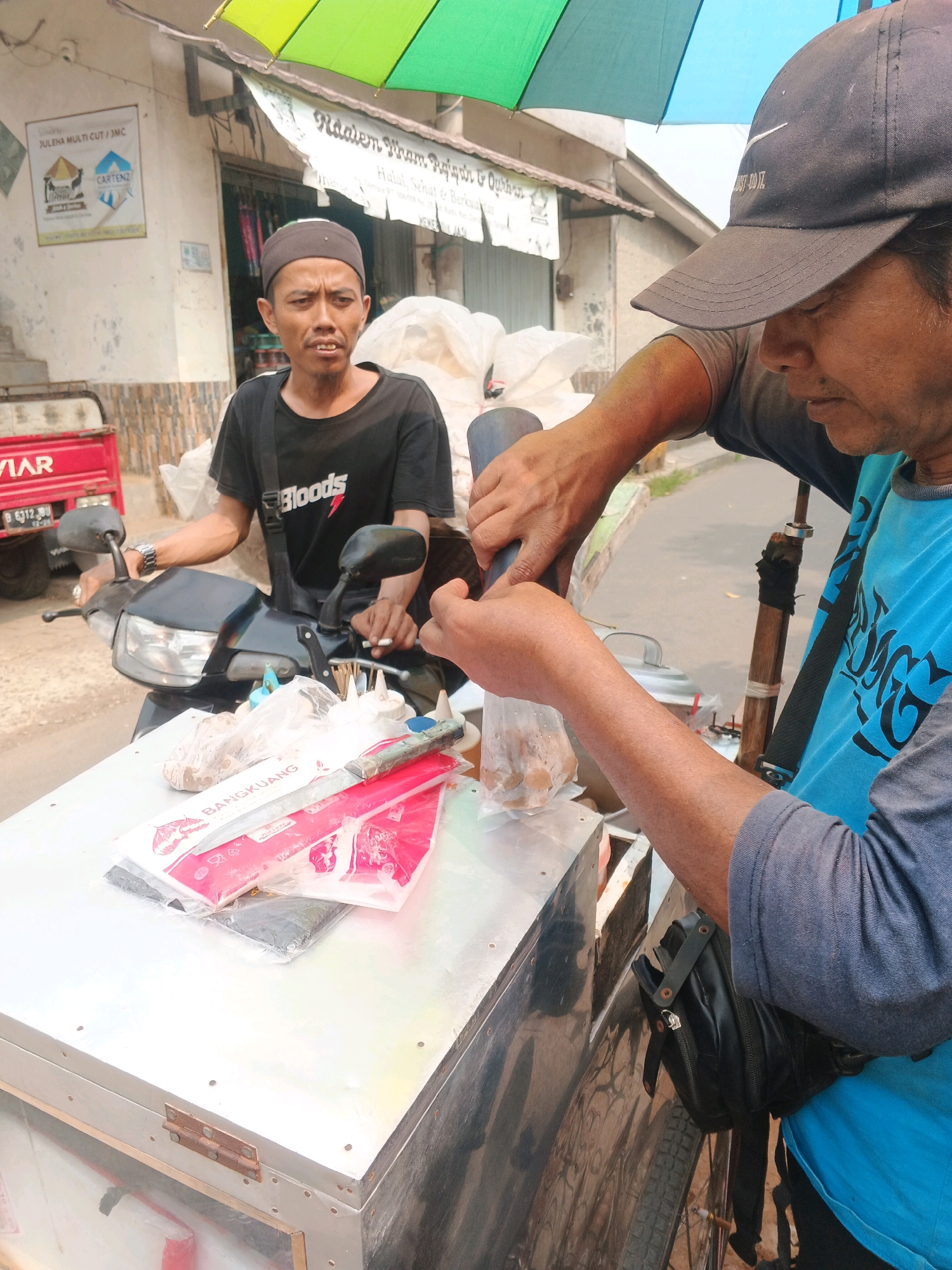 An outdoor market stall is depicted in Kadu, Indonesia. Two men are present. The man on the left, wearing a dark skullcap and a black t-shirt with white lettering, is seated on a motorcycle and appears to be looking to his right. The man on the right, wearing a dark baseball cap and a blue t-shirt, is standing and operating a food preparation device. He is holding a plastic bag containing food items with his left hand while using his right hand to guide a tube into the bag. In front of him is a metal cart with various items on top, including packaged goods, a knife, and skewers. Behind the men, a building with a sign that reads "JULEHA MULTI CUT/JMC" is visible. A multi-colored umbrella is partially visible overhead. The general environment suggests a street-side vendor setting.