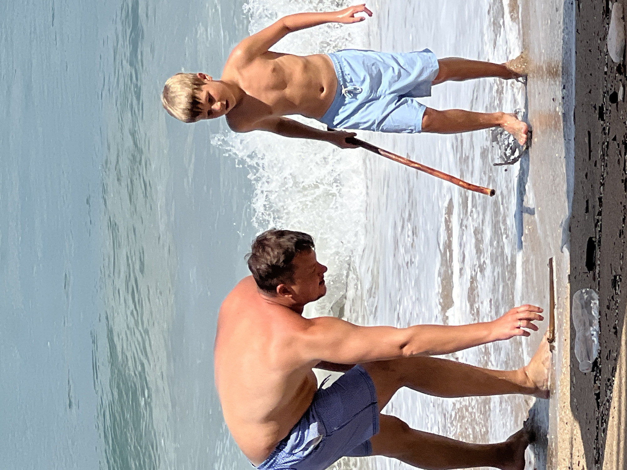 A father and son are on the beach in Kuta, Indonesia, by the sea. The father, wearing blue checkered swim trunks, is bent over near the waterline, reaching for something on the wet sand. The son, in light blue swim trunks, stands a few feet away holding a long wooden stick. The ocean water is calm with small waves crashing gently on the shore. There is debris on the sand in the foreground, including a plastic bottle.