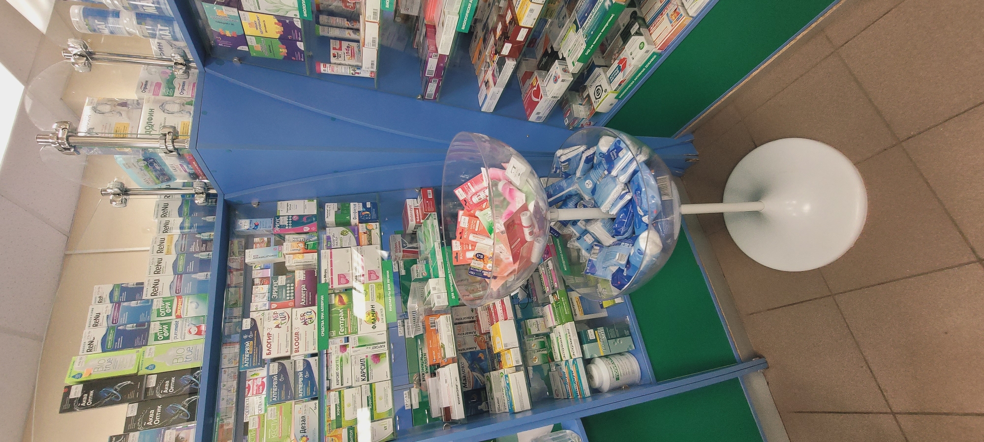 The image depicts the interior of a pharmacy, focusing on a display of medications and related products. The shelves are stocked with various boxes and containers, labeled with Cyrillic text. In the foreground, two transparent, spherical dispensers hold items, one filled with small, possibly cosmetic, products in red and pink packaging, and the other with blue and white wrapped items. These dispensers are mounted on a white stand. The shelves are blue and green. The floor is tiled in a neutral color. The scene gives the impression of a well-stocked, potentially Russian pharmacy.