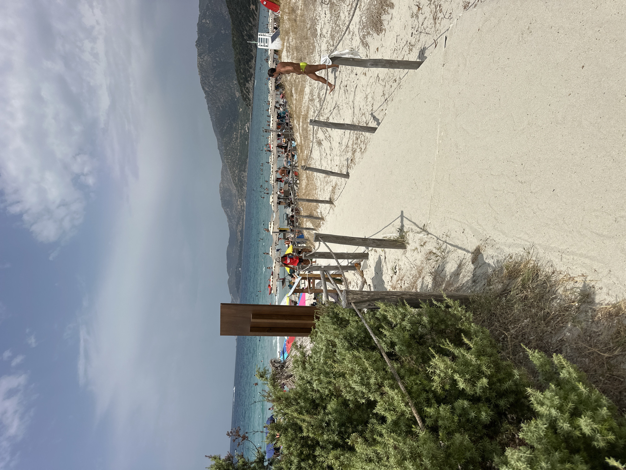 The image depicts a sandy beach scene in Crabonaxa, Villasimius, Italy. In the foreground, a sandy pathway, bordered by a rustic wooden fence with ropes, leads towards a crowded beach. Lush green bushes line the left side of the pathway. The beach is populated with numerous beachgoers relaxing under umbrellas and enjoying the turquoise water. In the middle ground, several colorful umbrellas are set up, and people are visible swimming and wading in the sea. A lifeguard stand is present on the right side of the beach. The background features a hilly coastline under a partly cloudy sky. A man in yellow swim trunks is walking on the pathway towards the beach in the right side of the image.