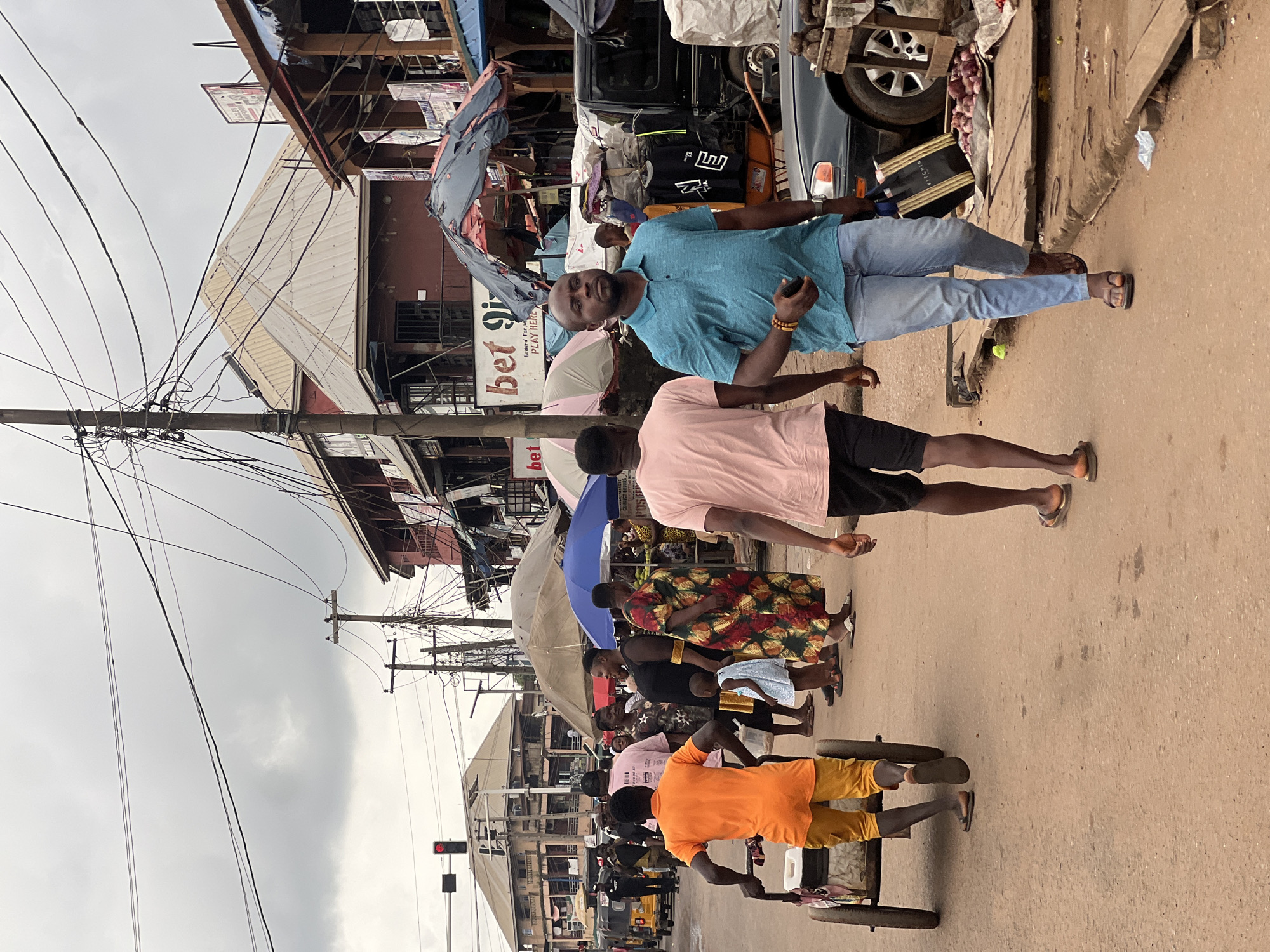 This image depicts a street scene in Nkpor, Nigeria, under an overcast sky. A man in a blue polo shirt and light blue jeans walks towards the viewer, carrying a dark shopping bag. To his left, a man in a pink t-shirt and dark shorts walks away from the viewer. In the foreground left, a man in an orange t-shirt and yellow shorts pushes a cart with two wheels. Behind him, a group of people are gathered, including a woman in a colorful patterned dress, a woman in a black dress, and a young child in a light blue dress. Further back, a yellow commercial tricycle is visible. The background is lined with buildings, some of which appear to be shops with signs and awnings. Overhead, a dense network of electrical wires is strung between utility poles, a common sight in urban Nigerian environments. The street is paved with dirt or asphalt. The overall impression is of a busy market or commercial area.