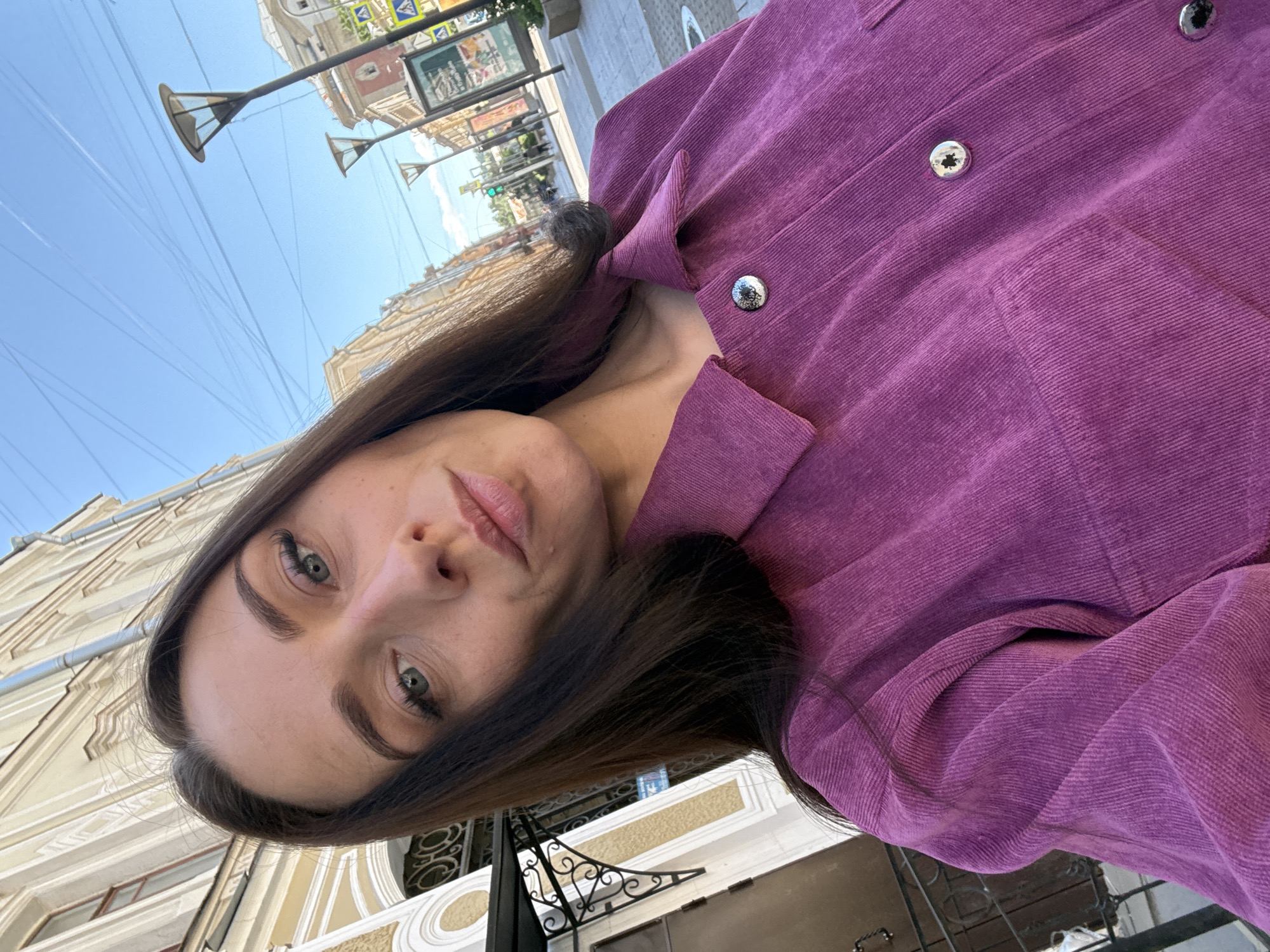 A woman with dark hair and blue eyes is taking a selfie outdoors on a street in Saint Petersburg, Russia. She is wearing a purple corduroy shirt. The background shows a street with buildings, lampposts, and power lines under a clear blue sky. The architecture suggests a historic district.