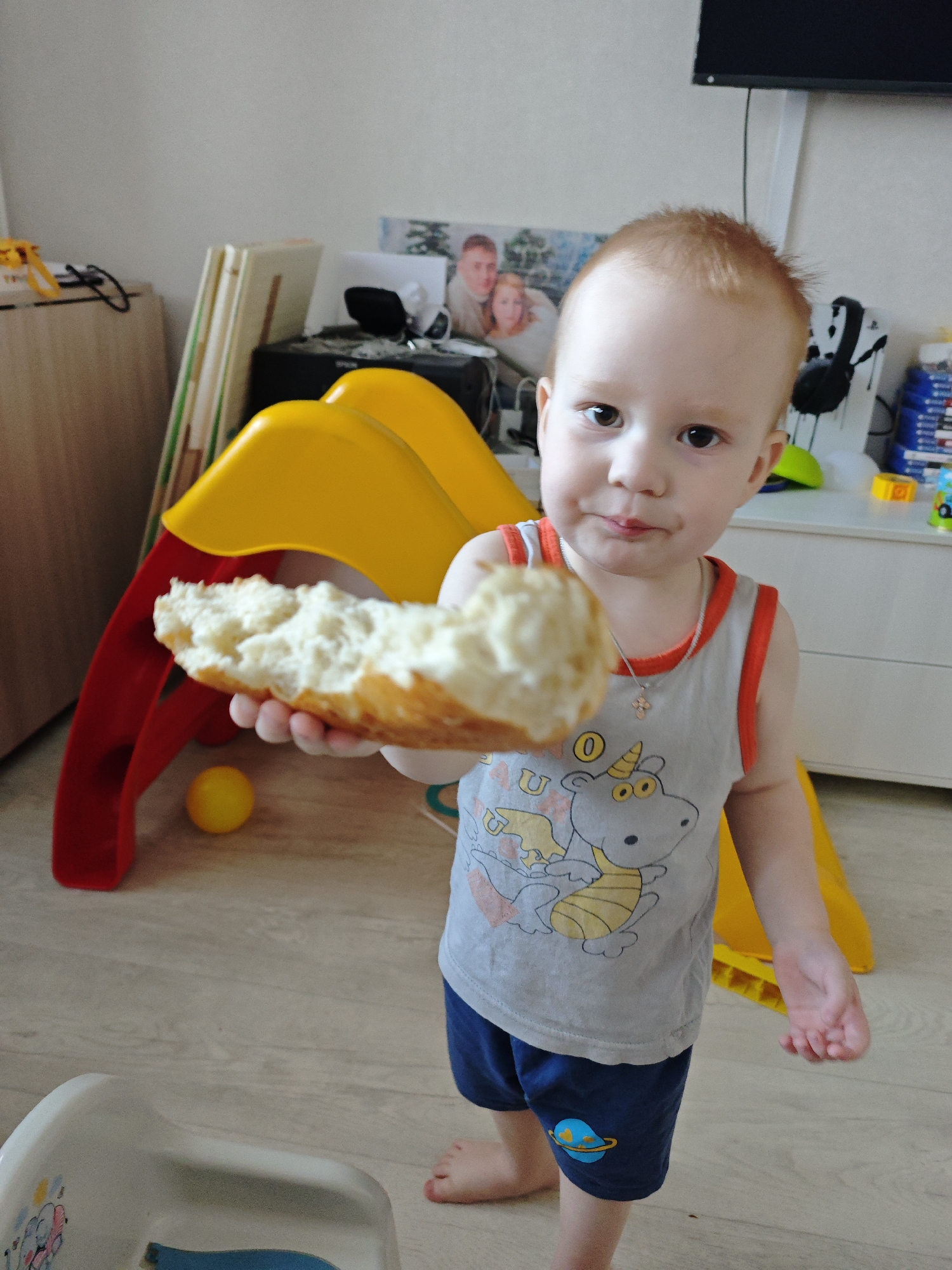 A young, fair-skinned child with reddish-blonde hair stands indoors holding a piece of bread. The child wears a grey tank top with a cartoon dinosaur design and navy blue shorts with a planet motif. A small silver cross hangs around the child's neck. The location is Markovskii, Russia, based on the prompt. The background includes a yellow and red plastic play structure, a light-colored wooden floor, a low white cabinet, and a stack of framed pictures. A television is mounted on the wall.