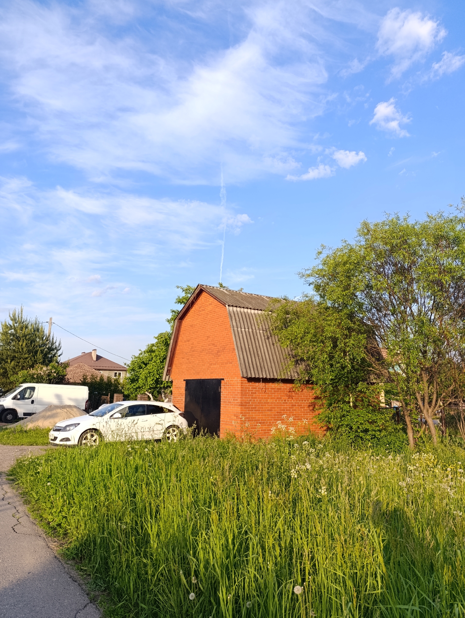The image captures a sunlit outdoor scene, likely a rural or suburban residential area, characterized by lush greenery and a clear sky. In the foreground, vibrant, tall green grass and wildflowers, including dandelions, stretch across the lower half of the frame. A narrow asphalt road or path, showing some cracks, runs along the bottom left edge.
Dominating the midground is a red brick building, possibly a barn, garage, or shed, featuring a dark grey corrugated roof and black double doors. To the left of this building, a white hatchback car is parked on the grass. Further left and slightly behind the car, a white cargo van is visible next to a pile of sand or gravel. A utility pole with power lines stands nearby.
Behind the red brick building, a large, leafy green tree with extensive foliage provides shade. In the distance, parts of other houses with brown roofs can be seen nestled among trees. The sky above is a bright, clear blue, adorned with scattered wispy white clouds and a faint, vertical airplane contrail in the upper right.
The lighting suggests late afternoon or early evening, with warm, golden sunlight casting long shadows and highlighting the textures of the grass and buildings. No people are visible, and no specific activity is taking place, conveying a tranquil and everyday atmosphere. There is no visible text.
