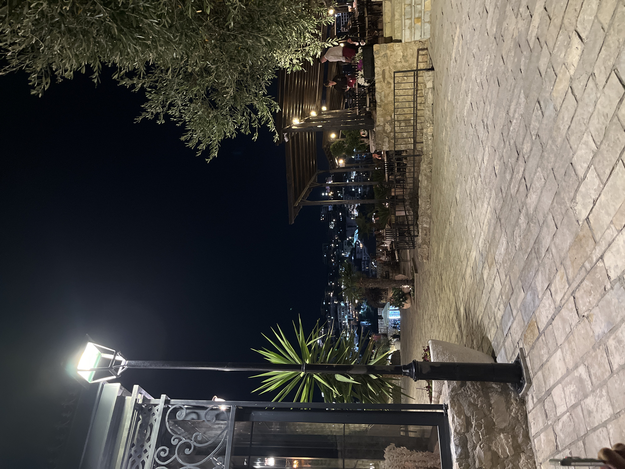 The image depicts a nighttime scene in the city of Ulcinj, Montenegro. A cobblestone pathway is illuminated by a tall lamppost on the left. In the background, a town with many lights is visible on a hillside. To the right, an outdoor seating area with tables and chairs is set up under a covered patio, with string lights providing illumination. Two servers, one in a white shirt and red apron and the other in a black shirt and red apron, are visible in the seating area. An olive tree with its branches extending into the frame is on the far right. A large yucca plant stands near the lamppost. The overall atmosphere suggests a restaurant or cafe operating in the evening.