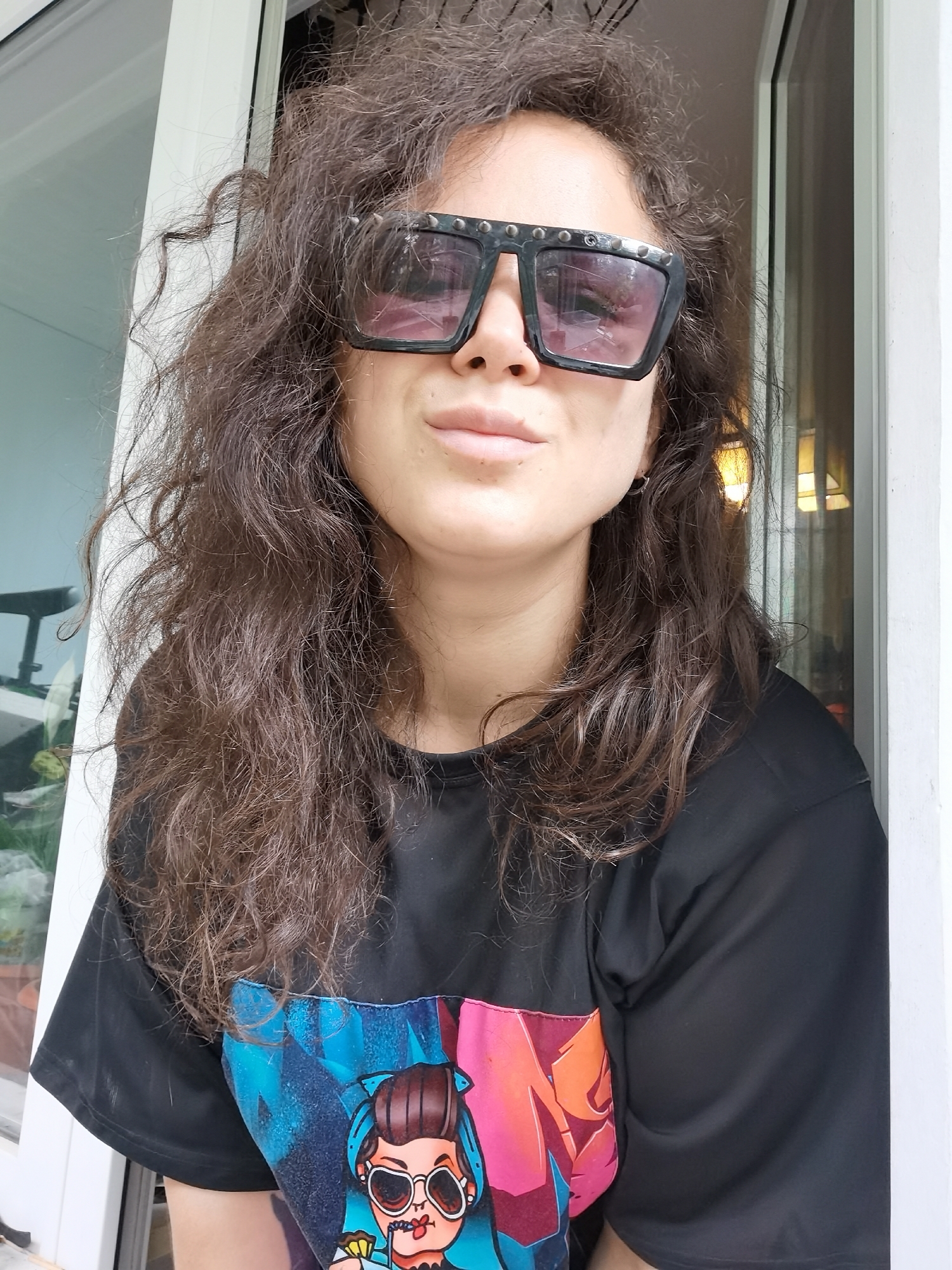 The image displays a head and torso shot of a person, likely female, looking directly at the camera. She has voluminous, dark brown, curly hair that appears somewhat disheveled and frames her face.
The person is wearing large, square-framed sunglasses with tinted purple-pink lenses and silver stud embellishments along the top edges of the frames. Reflections visible in the lenses suggest bright ambient light and potentially interior elements. Her lips are slightly parted in a neutral expression, and she has visible pores and minor skin texture. A small, silver hoop earring is visible on her right earlobe.
She is dressed in a black short-sleeved t-shirt featuring a prominent, colorful graphic design on the chest. The graphic depicts a stylized cartoon character, a woman with brown hair styled in a bun, wearing large sunglasses, and holding a straw or lollipop in her mouth. The background of the graphic consists of vibrant, abstract geometric shapes in shades of blue, pink, and orange.
The background of the shot is bright, suggesting natural light, likely daytime. White door or window frames are visible on both sides of the person, indicating she is positioned near an opening. Reflections in the background glass panels show blurry shapes that could be indoor objects or distant outdoor elements. No explicit text is visible, only the cartoon graphic on the t-shirt. The person appears relaxed and is posing for the photograph.