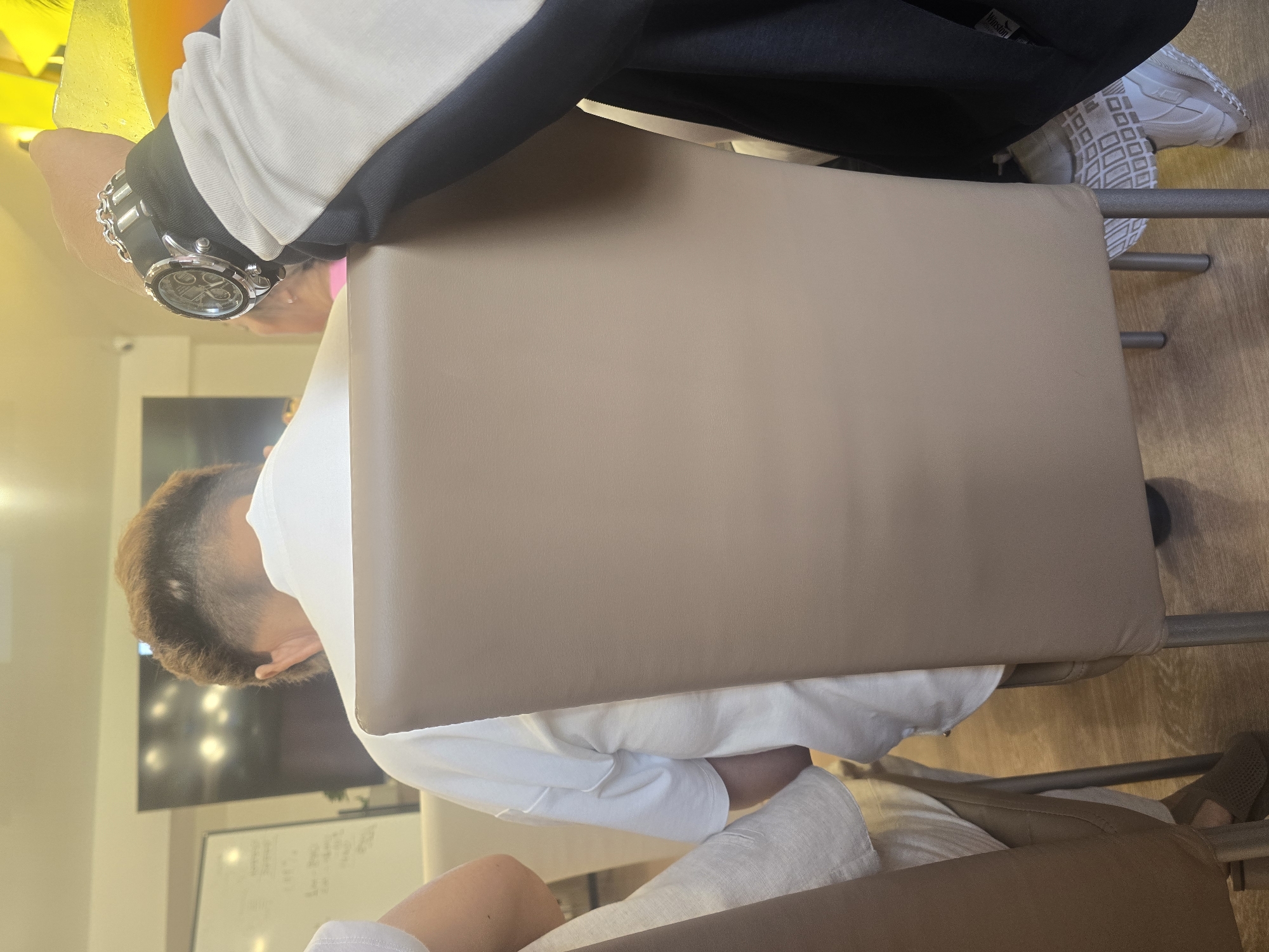 The image shows the back of a person wearing a white t-shirt, seated on a tan chair. The person has short, styled brown hair. To the right of this person, another individual's arm is visible, wearing a watch with a black band and silver detailing. This individual is also wearing a dark jacket over a white sleeve. In the background, a whiteboard with writing and a television screen with lights are visible. The setting appears to be indoors, possibly a cafe or meeting room, with additional chairs and people partially visible. The image does not provide any specific information to indicate the location is Ulan-Ude, Russia.