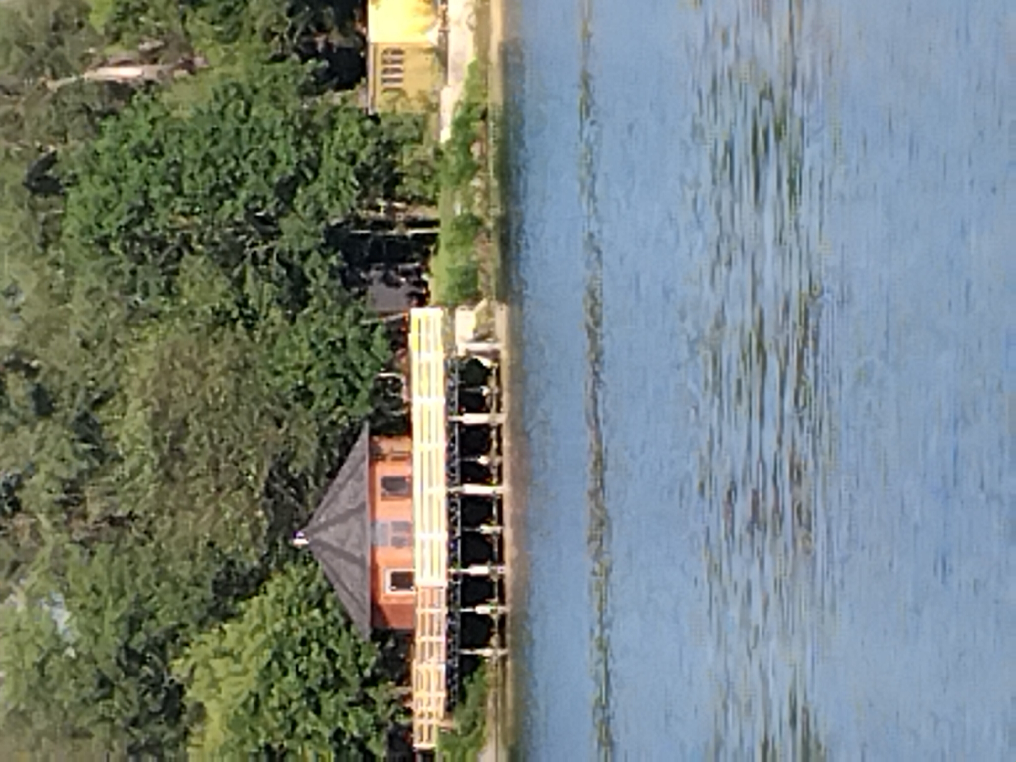 A lakeside scene features a multi-tiered structure with a small, orange-colored building at the top, supported by a wooden pier. The building has a dark, conical roof and several windows. A wooden railing runs along the pier. The structure is situated on the shore, backed by dense, green foliage and trees. The water in the foreground is a vibrant blue, reflecting the sky and the surrounding greenery with rippling patterns. In the background, a yellow building with arched windows is partially visible. The scene is serene and appears to be a recreational area.